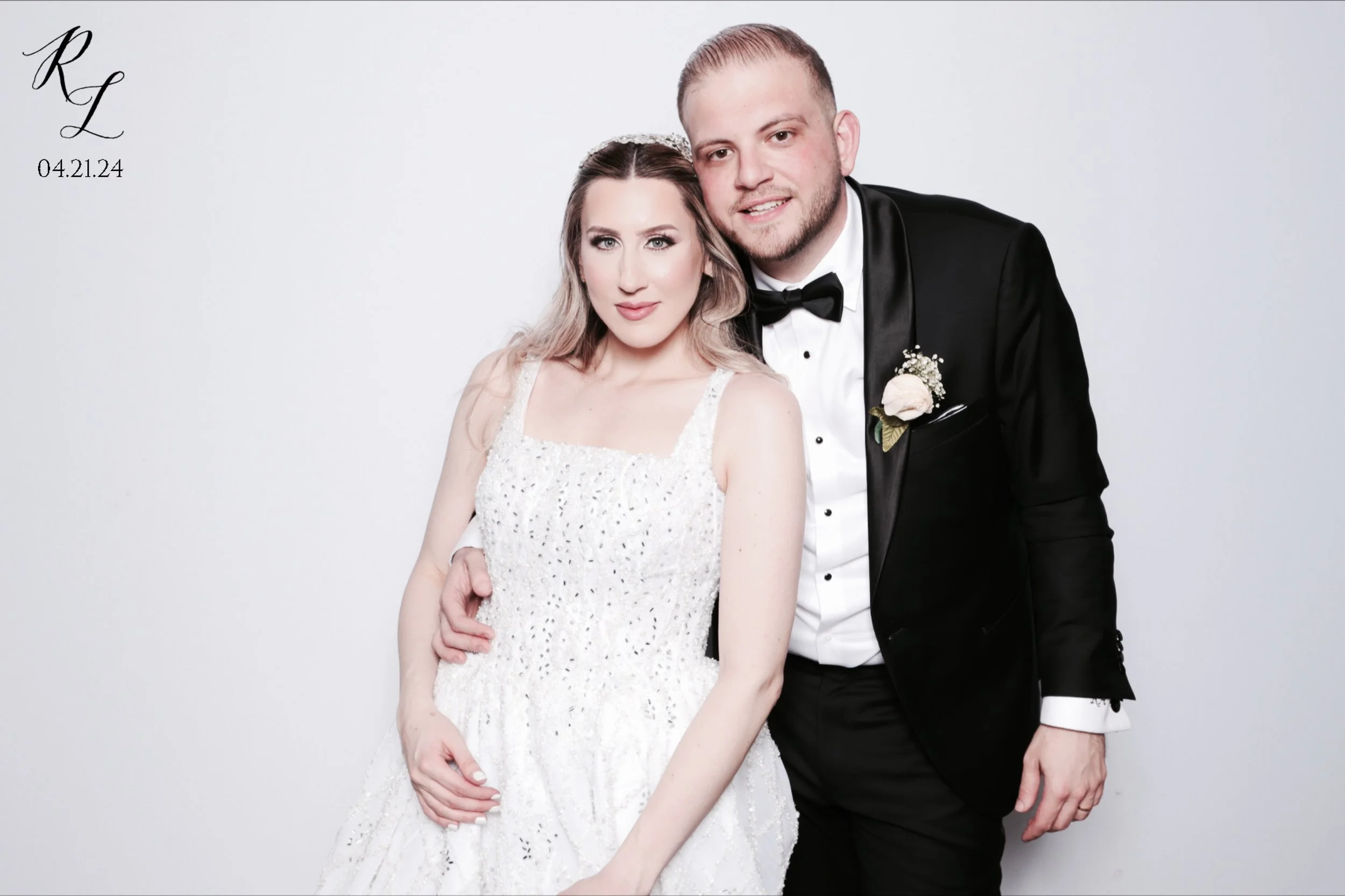 A bride and groom in wedding attire posing together against a plain white background. The bride is wearing a white lace dress and the groom is in a black tuxedo with a bow tie.