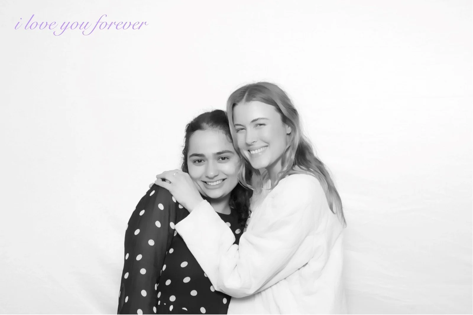 Two women hugging and smiling in front of a plain background, with purple text in the top left corner that says 'i love you forever'.