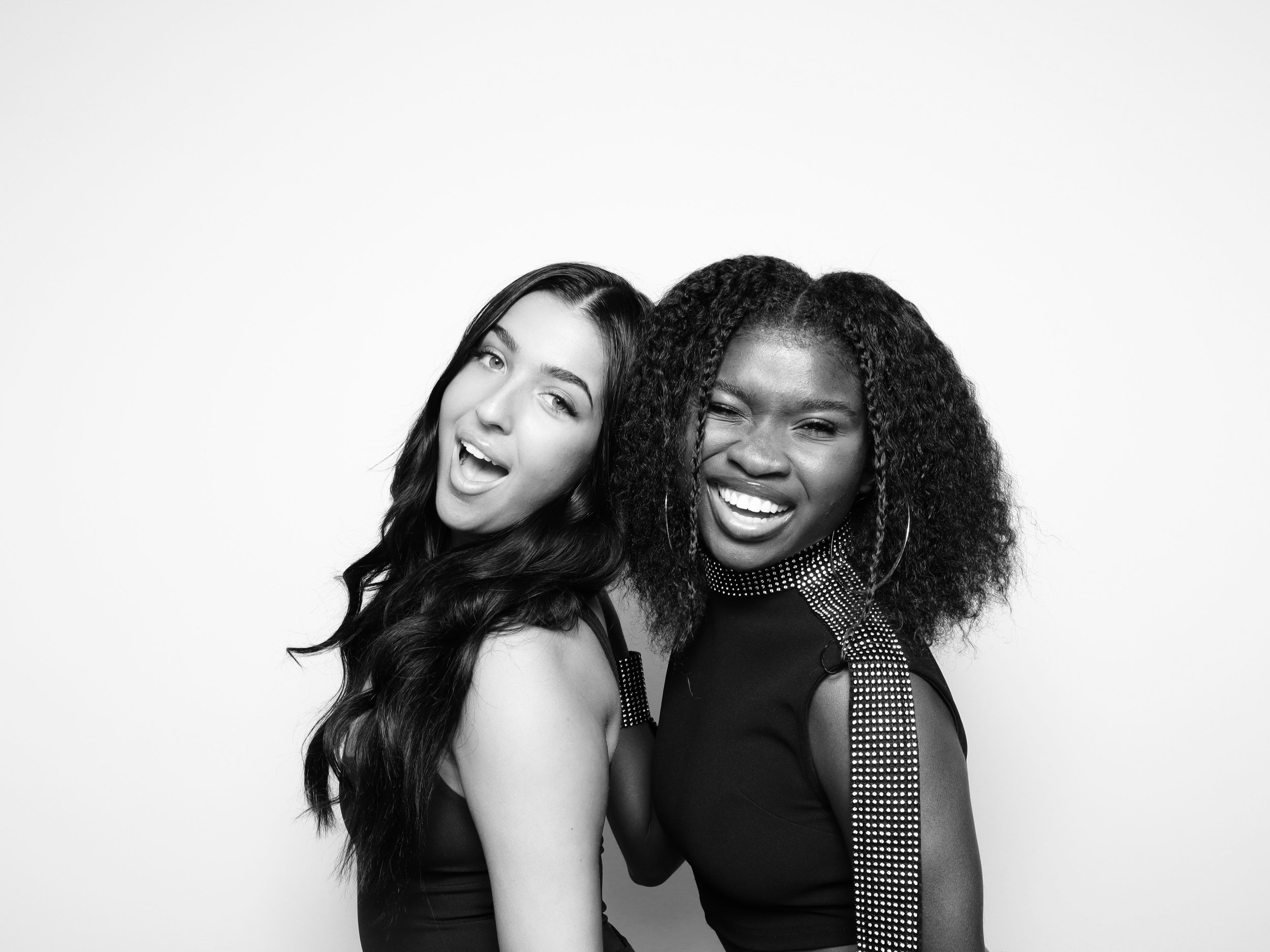 Two women smiling and posing together in black outfits, one with long dark hair and the other with curly hair, against a glam background.