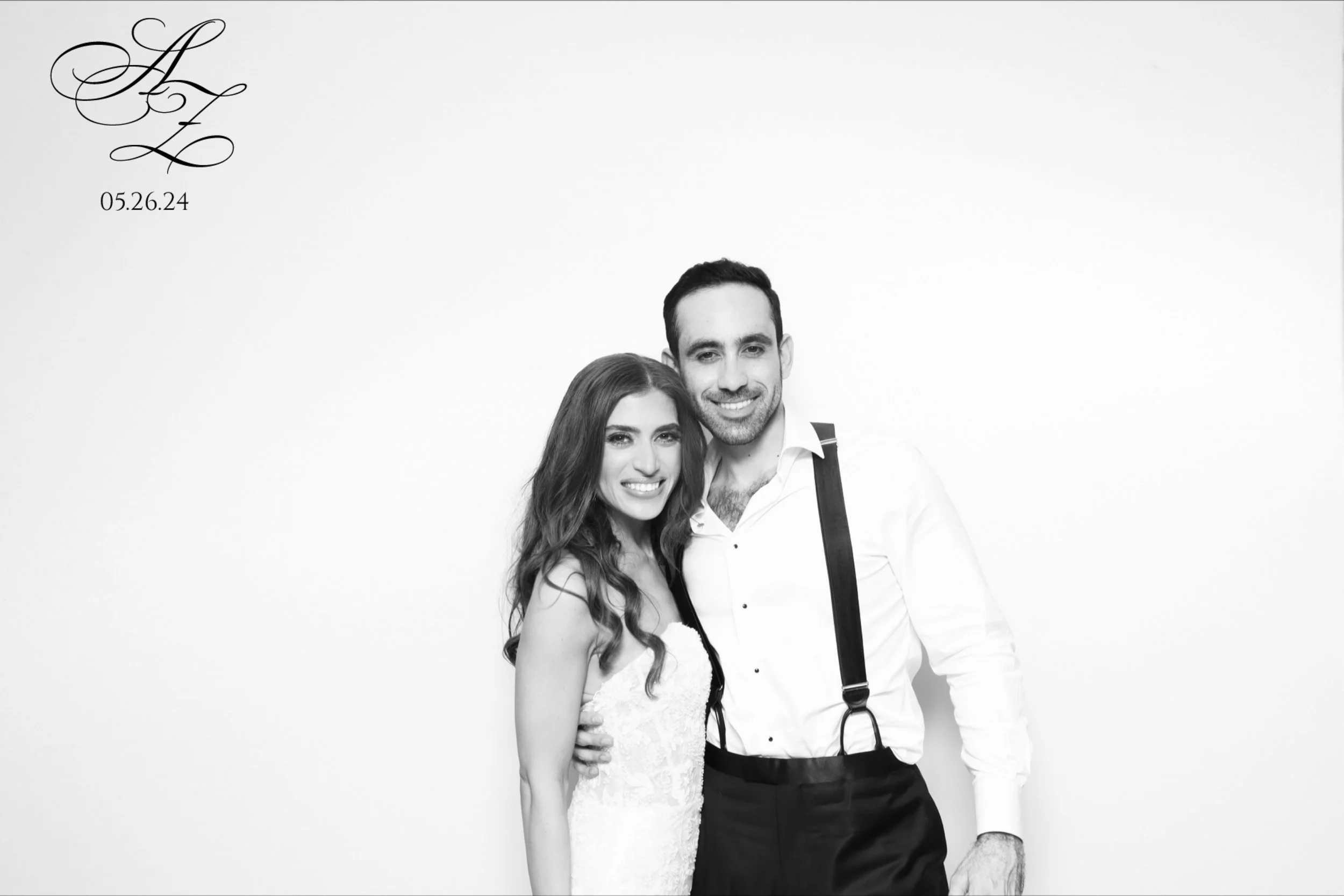 A smiling couple dressed in formal attire standing close together, with the woman wearing a lace dress and the man wearing a white shirt with suspenders, against a plain white background. There is a monogram or logo with initials in the top left corn