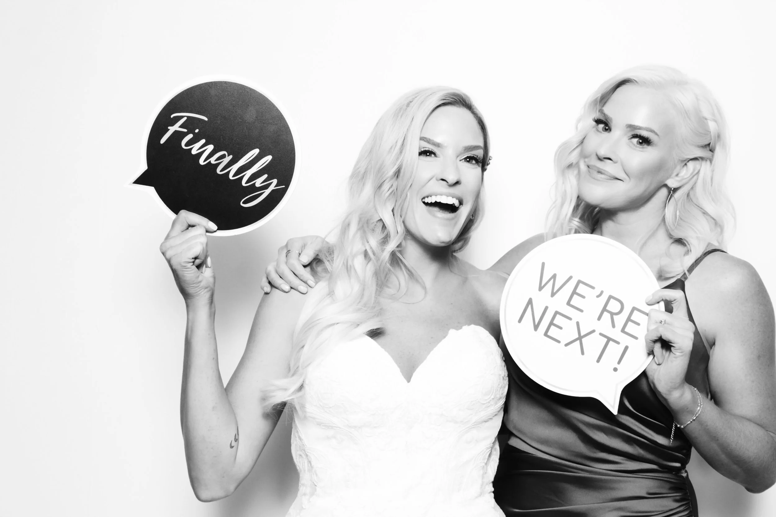 Two women at a wedding celebration holding speech bubble signs, one says 'Finally' and the other says 'We're Next!'