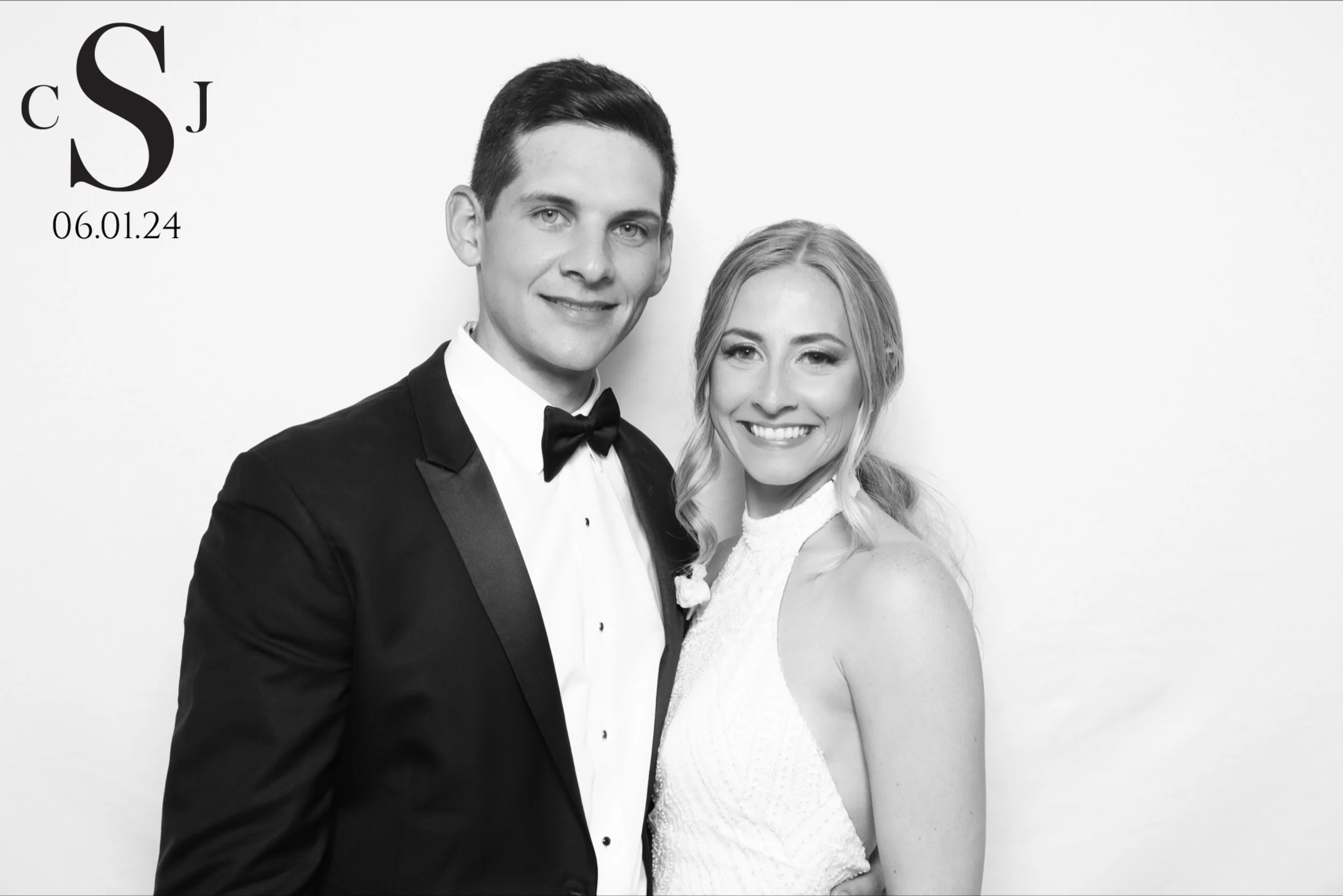 A black and white photo of a smiling couple in formal attire, with the man wearing a tuxedo and the woman wearing a white dress. There are initials and a date in the top left corner.