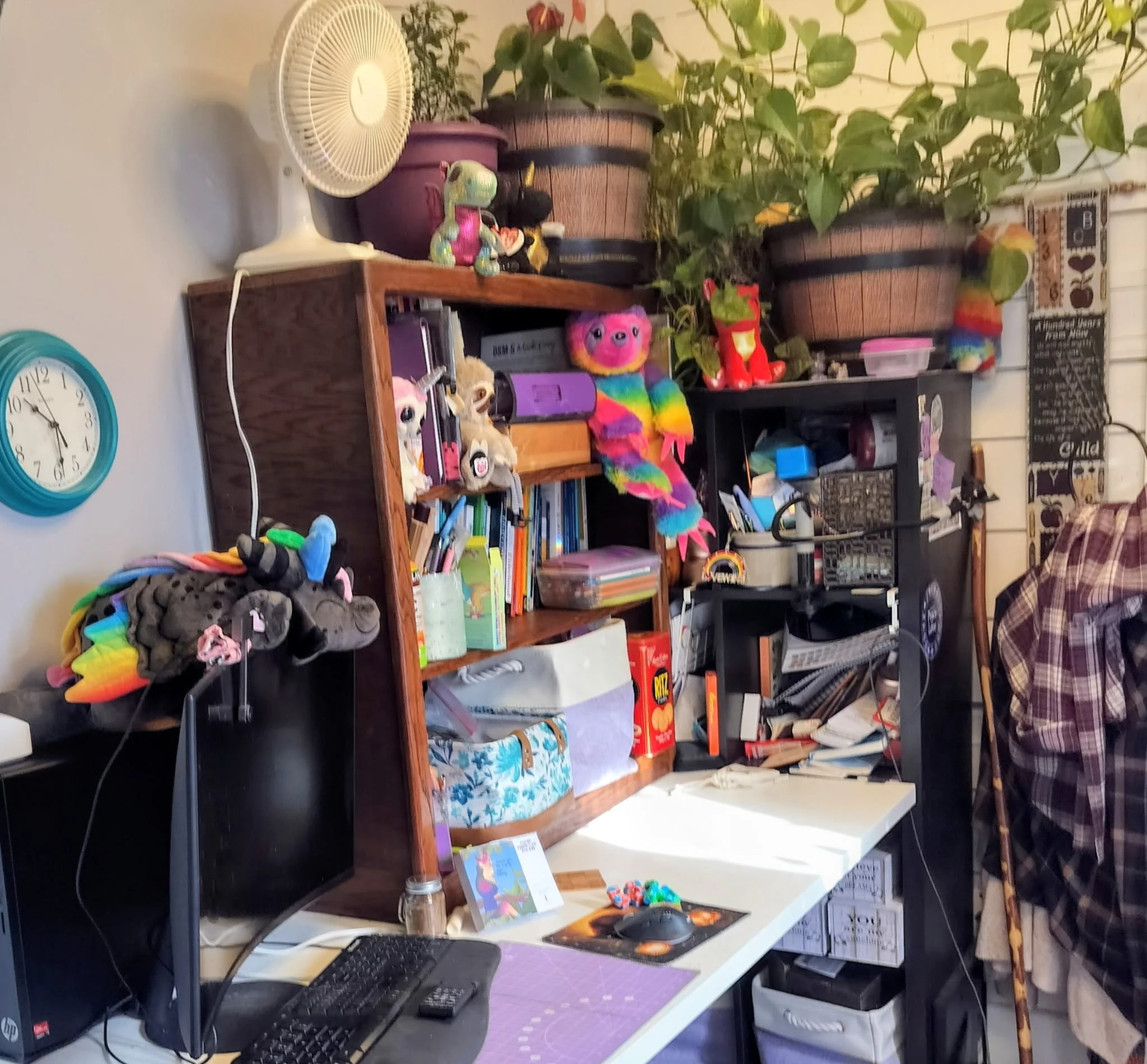 An organized workspace with a black computer and a purple mouse pad on a white desk. Behind the desk, there are wooden shelves filled with books, toys, and various knick-knacks. A fan is on the wall to the left, and a wall clock showing approximately 8:50 is also visible. The background includes potted plants and a chalkboard with writings and drawings.