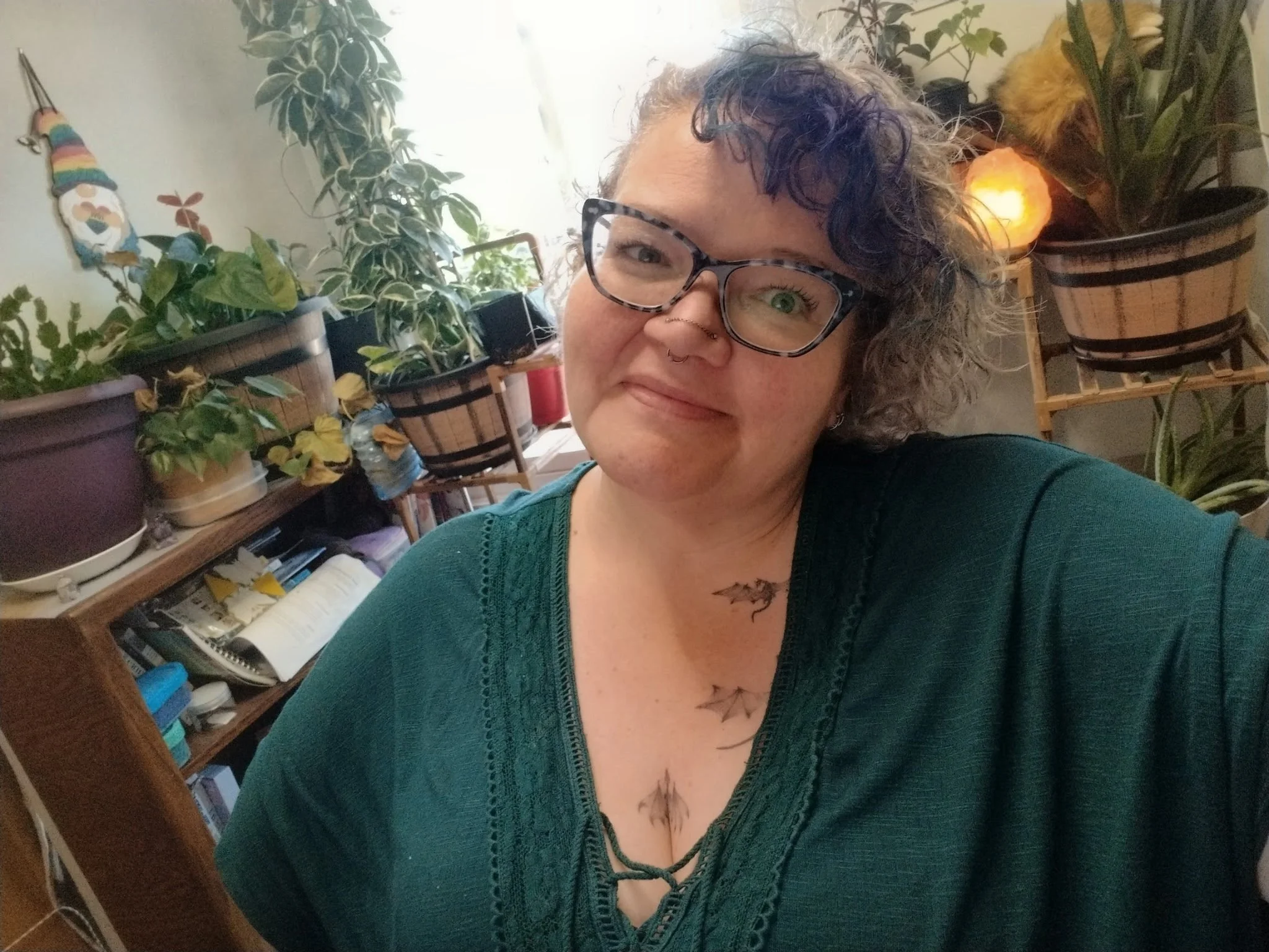 A woman with curly hair, glasses, and a septum piercing smiling in front of shelves with potted plants and books, with a salt lamp in the background.