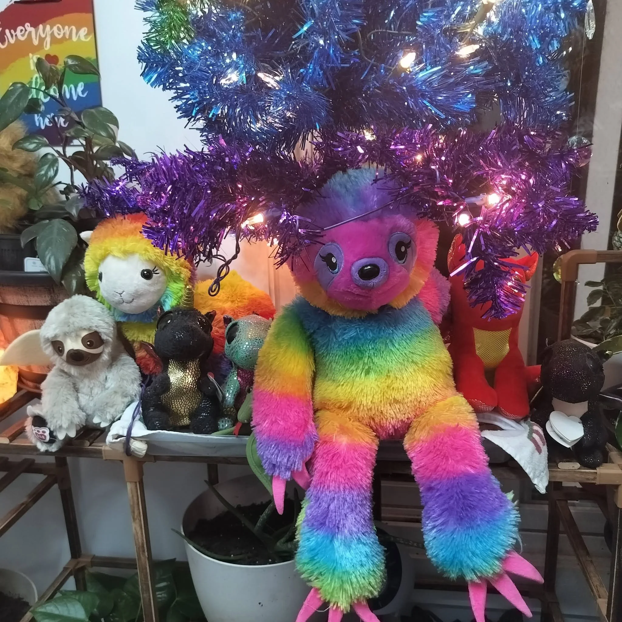 Colorful rainbow-themed plush lion toys with rainbow-colored fur and rainbow pom-pom tails, displayed on a table along with other plush toys, next to small potted plants and a rainbow-colored sign that reads 'Everyone is Welcome'.