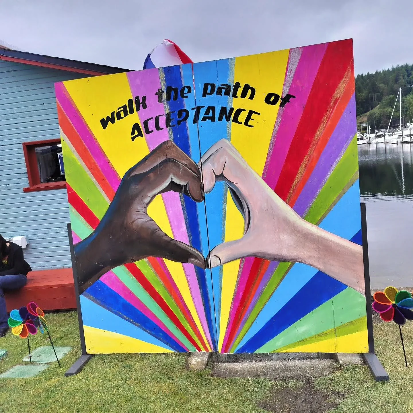 Colorful artwork with two hands forming a heart, Rainbow rays background, and the phrase 'walk the path of acceptance' at the top, outdoors near water and boats.