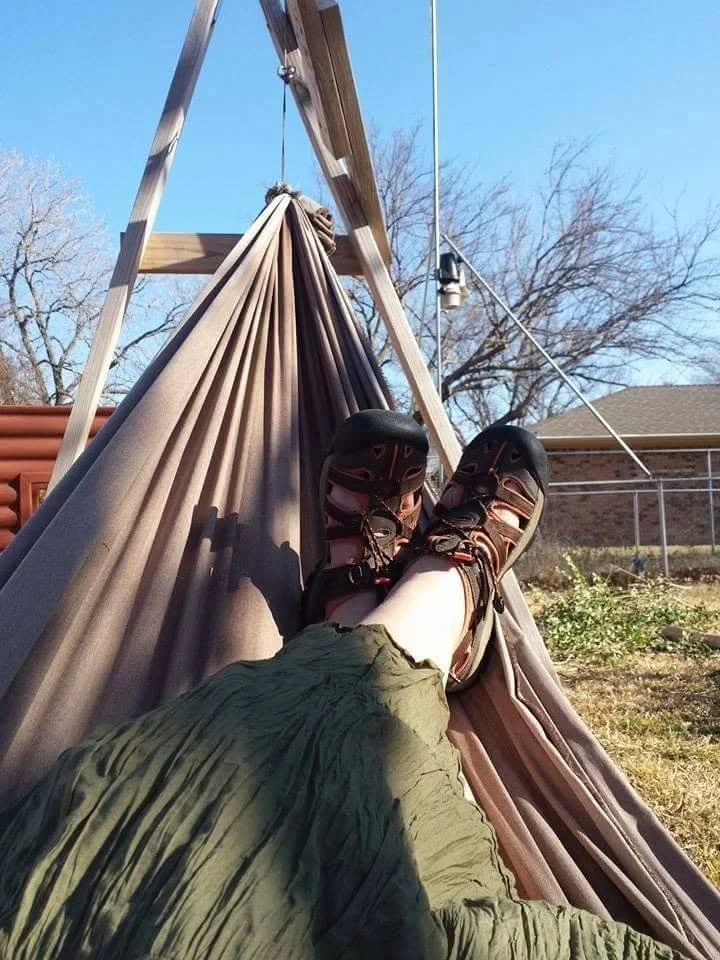 Person's legs and feet in outdoor sandals resting on a hammock tied to a wooden stand, with trees, a house, and a clear blue sky in the background.