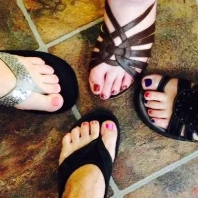 Four people standing in a circle, showing their feet in sandals with painted toenails on a tiled floor.