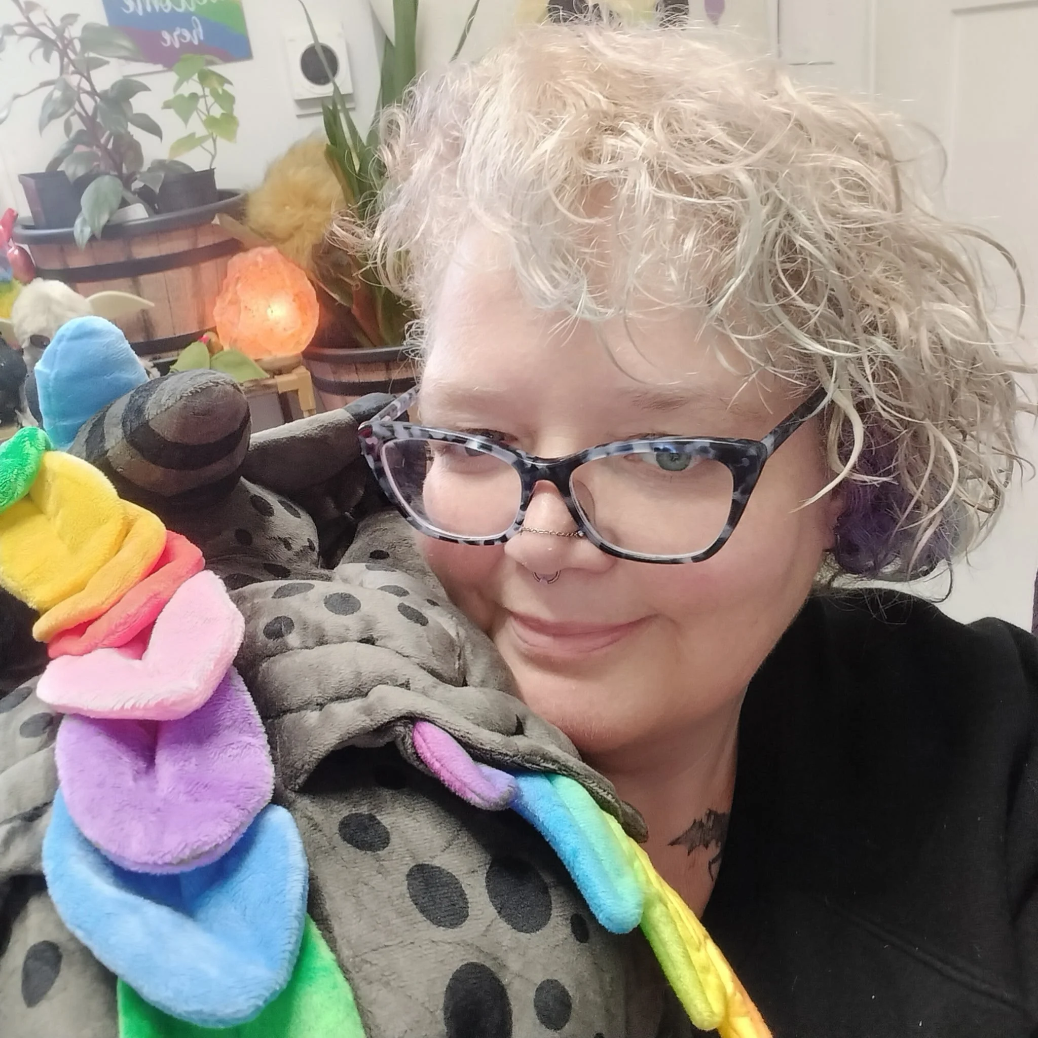 Close-up of a woman with curly blonde hair and glasses, wearing a rainbow-colored fuzzy blanket, surrounded by plush toys and a rainbow-themed blanket. Background includes plants, a salt lamp, and colorful decorations.