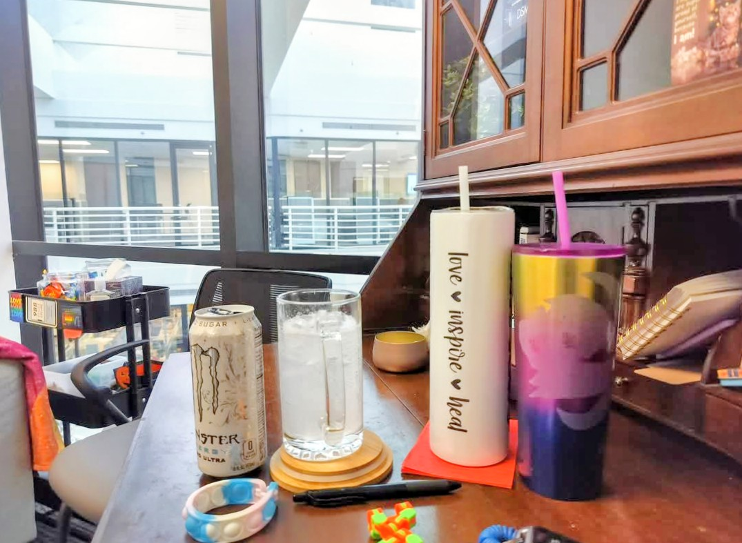 A wooden counter with a water glass, a Monster energy drink can, two large colorful cups with straws, a small white bowl, a black marker, and a multicolored bracelet. In the background, there is a window with a view of the outside, and a shelf with various small items.