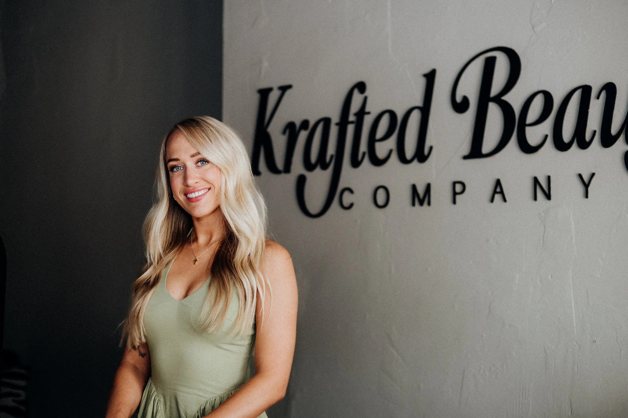 Brianna Wilderman in a light green dress stands indoors next to a wall with the words "Krafted Beauty Company" written on it.