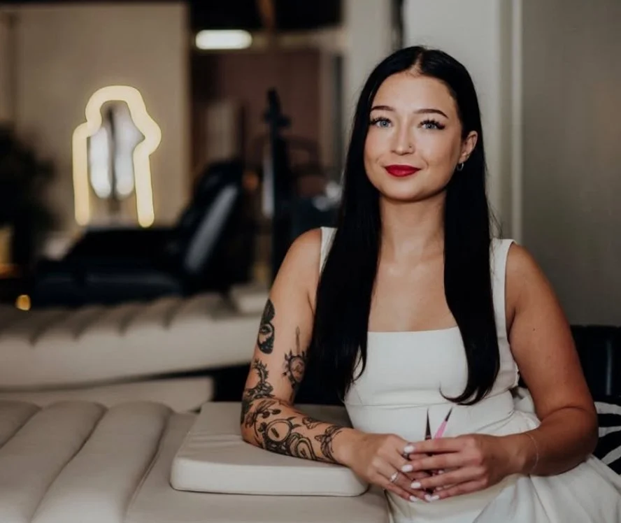 Emma Steinick sitting on a cream-colored sofa in a modern interior. She is wearing a white sleeveless dress and holding a pink card, smiling slightly at the camera.