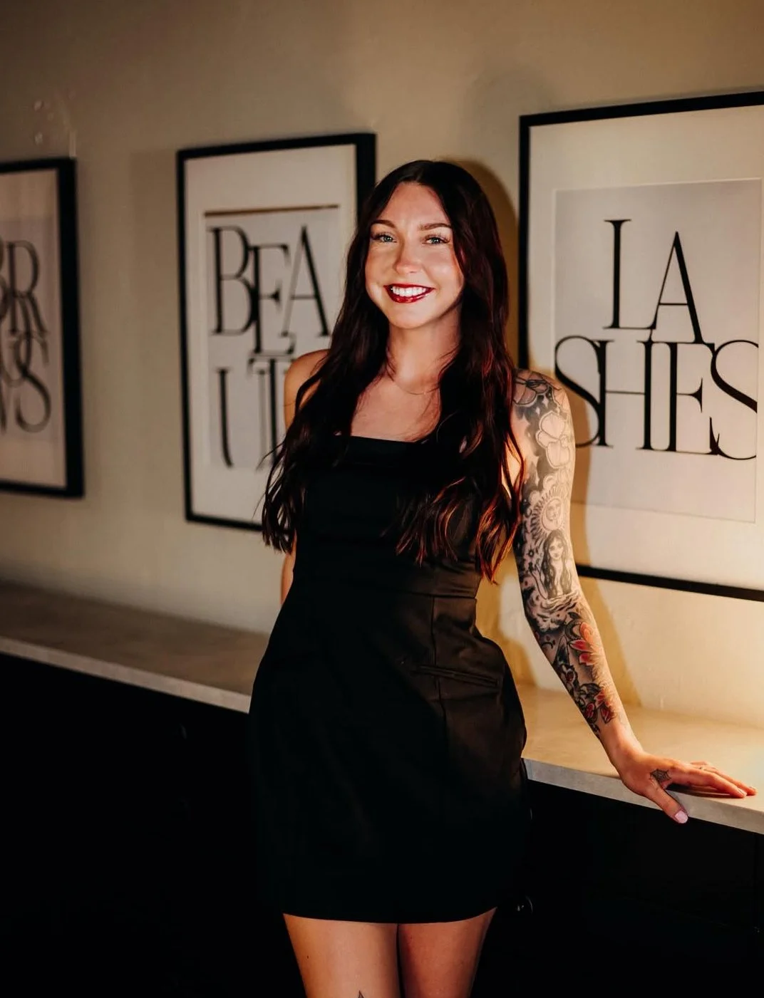 Ellie Braafhart, smiling and wearing a black dress, standing in front of framed wall art with typography phrases.