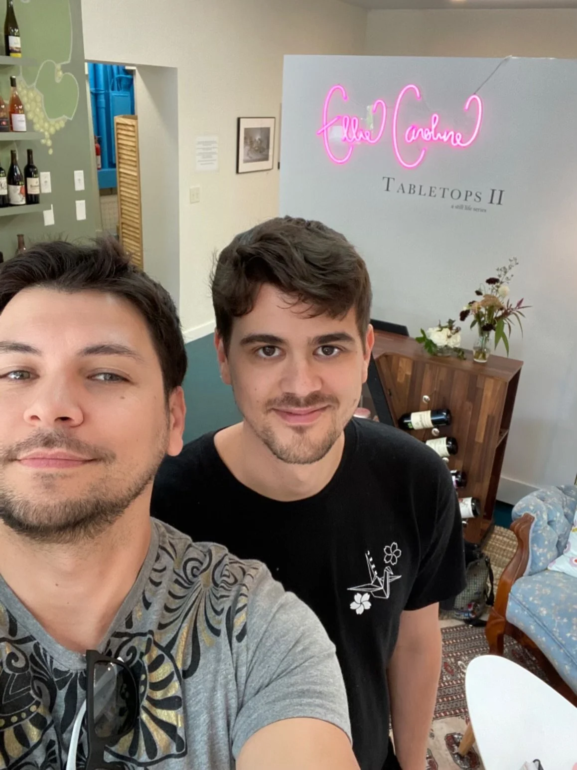 Two people taking a selfie in a room with wine bottles on shelves, a neon sign reading "Ellie Caroline Tabletop II," and a floral arrangement on a wooden table.