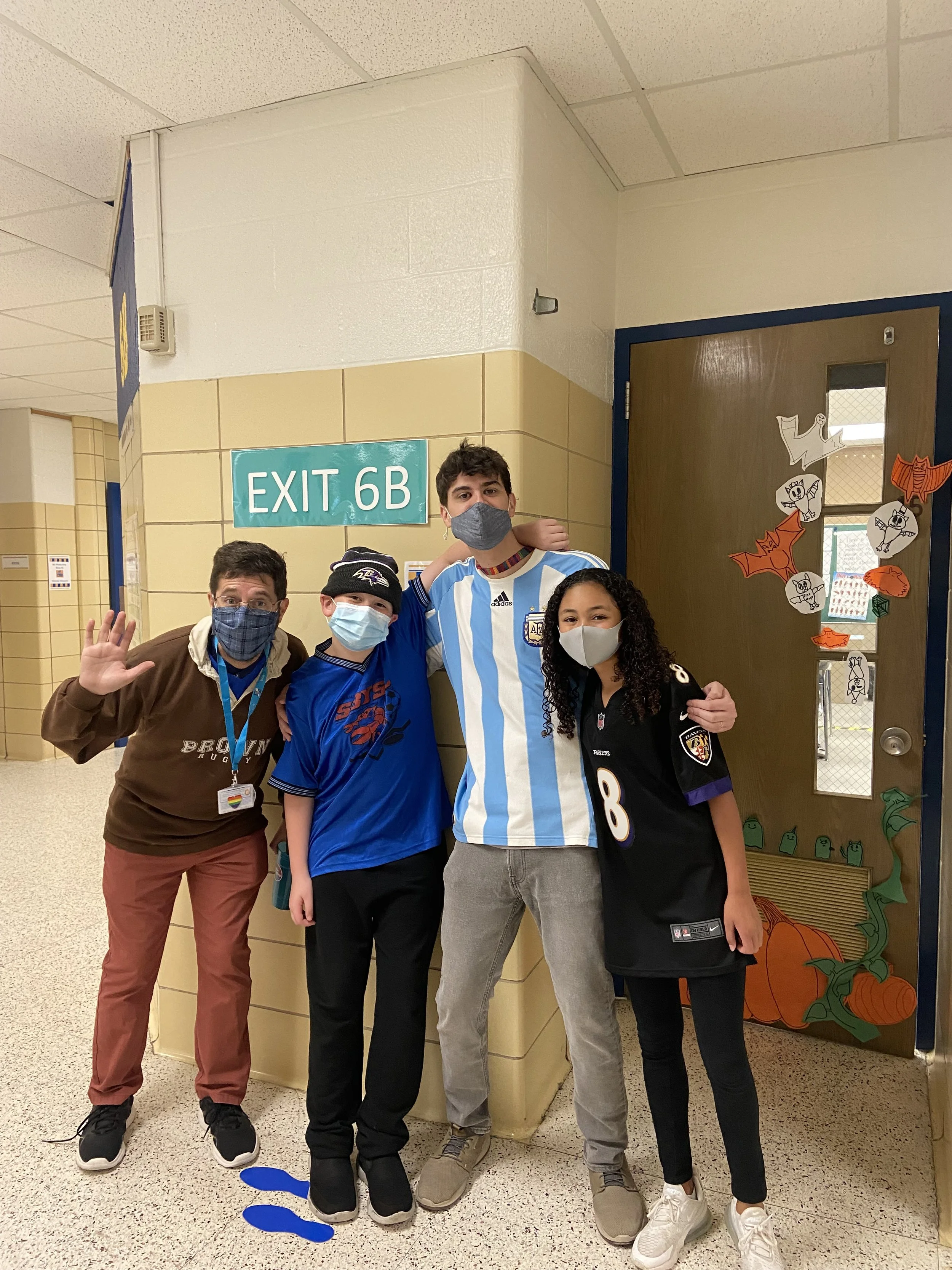 Group of four people in a hallway wearing masks, with exit sign 6B and decorated door in background.