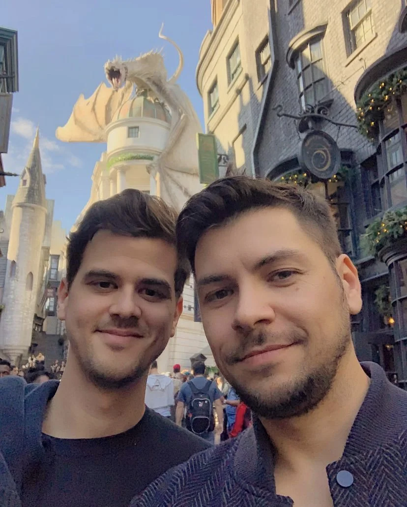 Two men taking a selfie in a theme park with a dragon atop a building.
