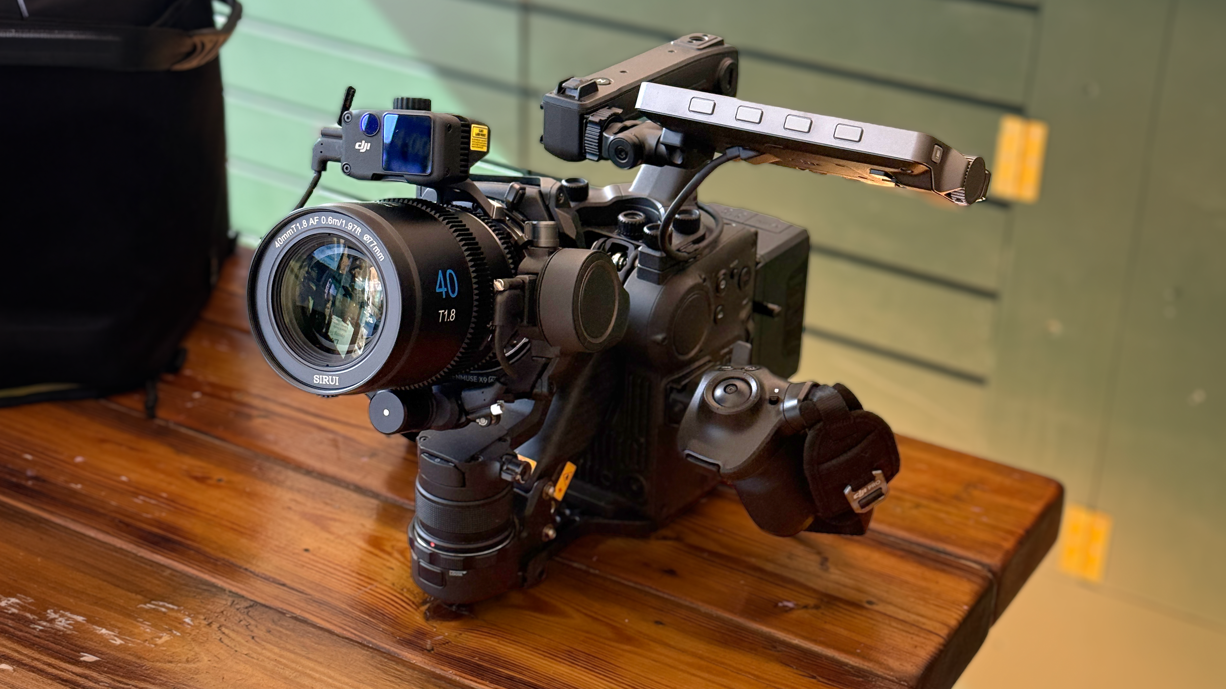 A professional video camera mounted on a tripod on a wooden surface, with additional camera equipment attached, and a blurred green wall background.