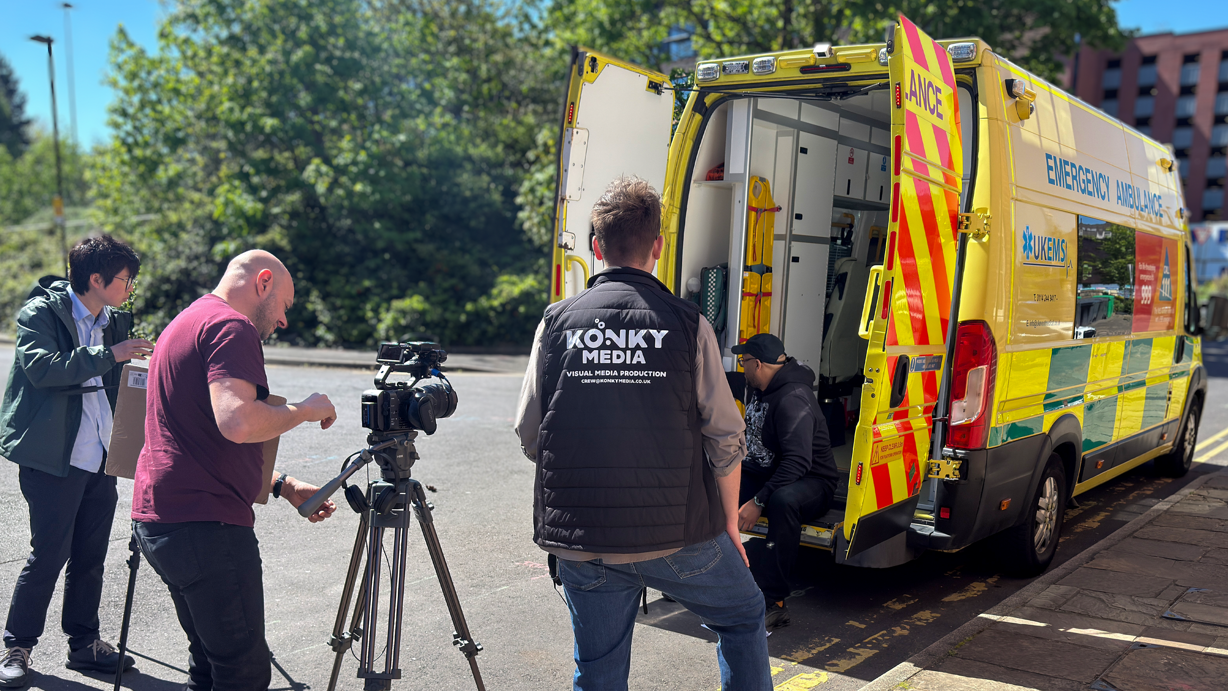A group of people filming with a camera and tripod near an emergency ambulance with open rear doors on a street with green trees and a multi-story building in the background.