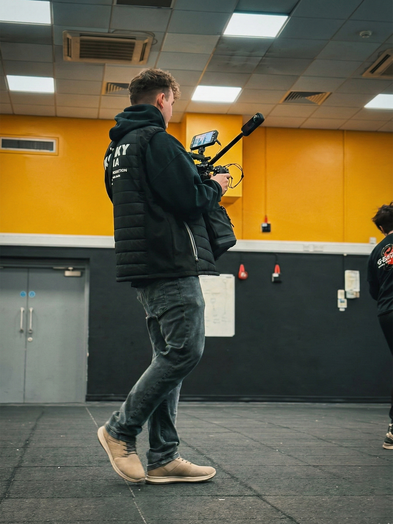 A young man standing indoors, holding a camera drone with an attached mobile device, wearing a black hoodie and jeans in a room with yellow walls and a black floor.
