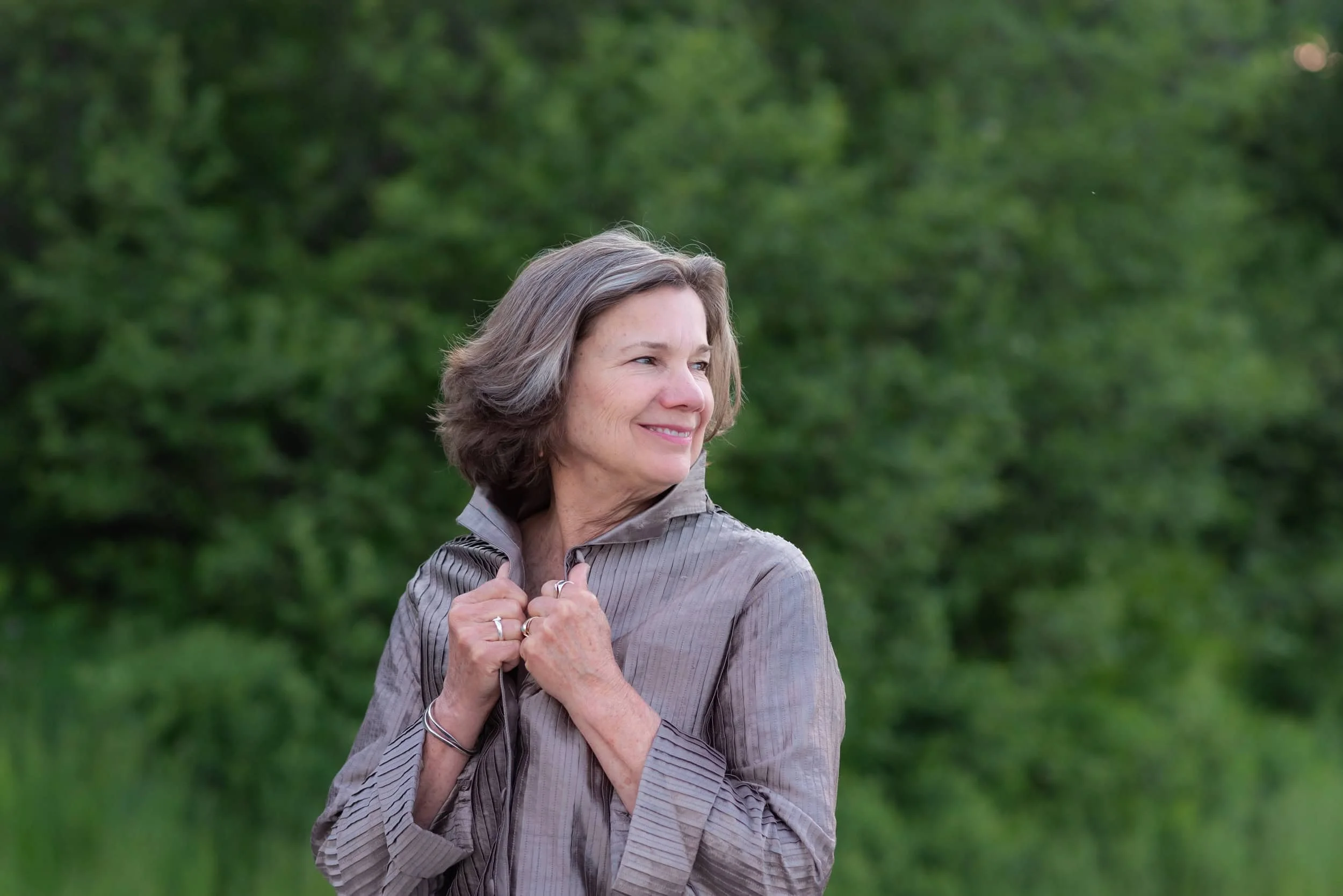 An woman with short gray hair wearing a blue sleeveless dress standing outdoors in a green, wooded area.