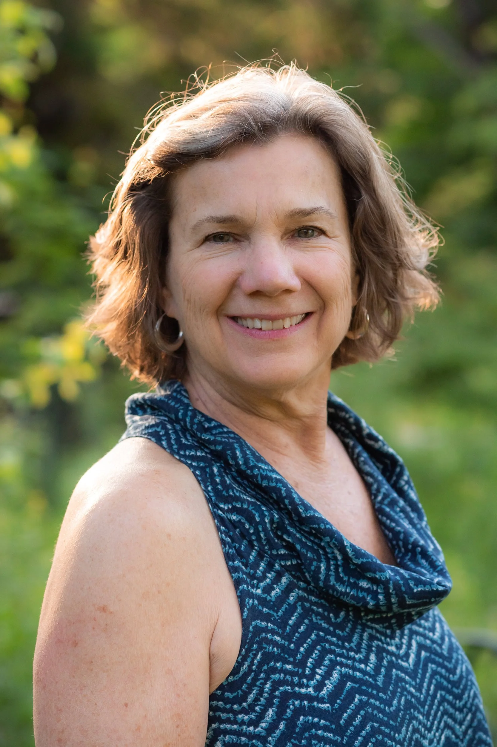 A smiling woman with short, curly brown hair, wearing a sleeveless, patterned blue top, standing outdoors against a blurred green background.