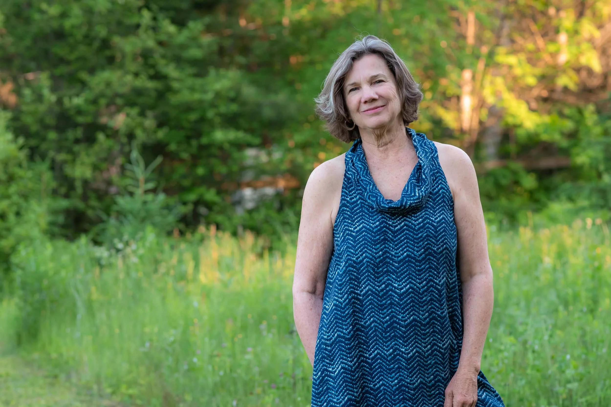 A woman with shoulder-length gray hair wearing a sleeveless blue patterned dress standing outdoors in a lush green setting with trees and grass.