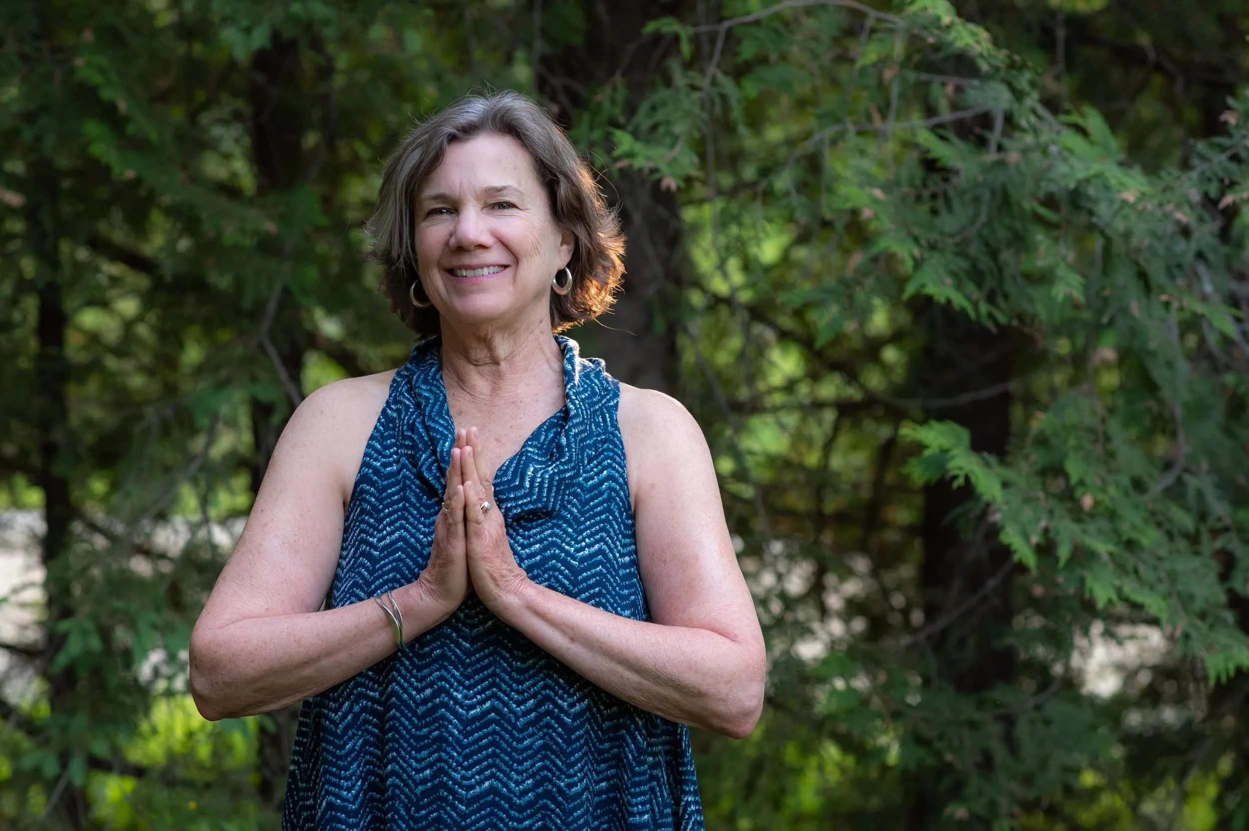 Woman smiling outdoors with her hands in a prayer position, surrounded by green trees and foliage.