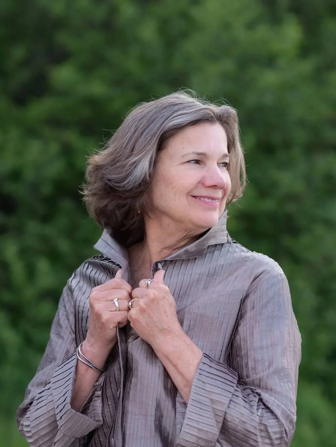 A middle-aged woman with shoulder-length gray hair smiling outdoors, holding the collar of her gray jacket against a blurred green background.