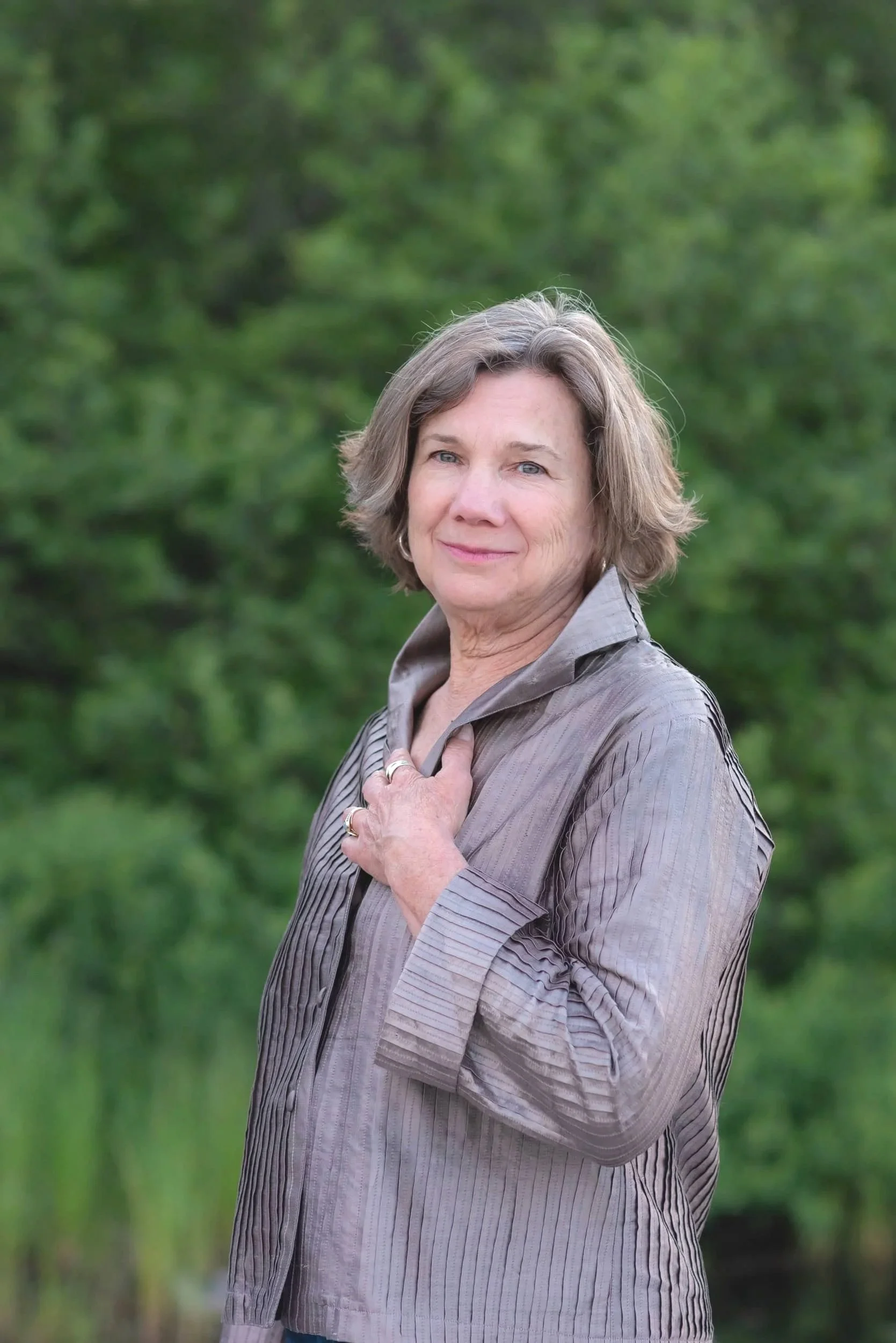 A woman with short gray hair standing outdoors in front of green trees and bushes, wearing a shiny gray blouse, looking at the camera with a gentle smile.