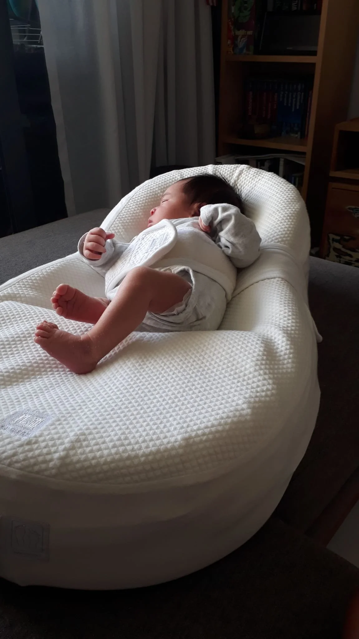A sleeping baby lying on a white cushioned changing pad, resting on a gray surface in a home setting.