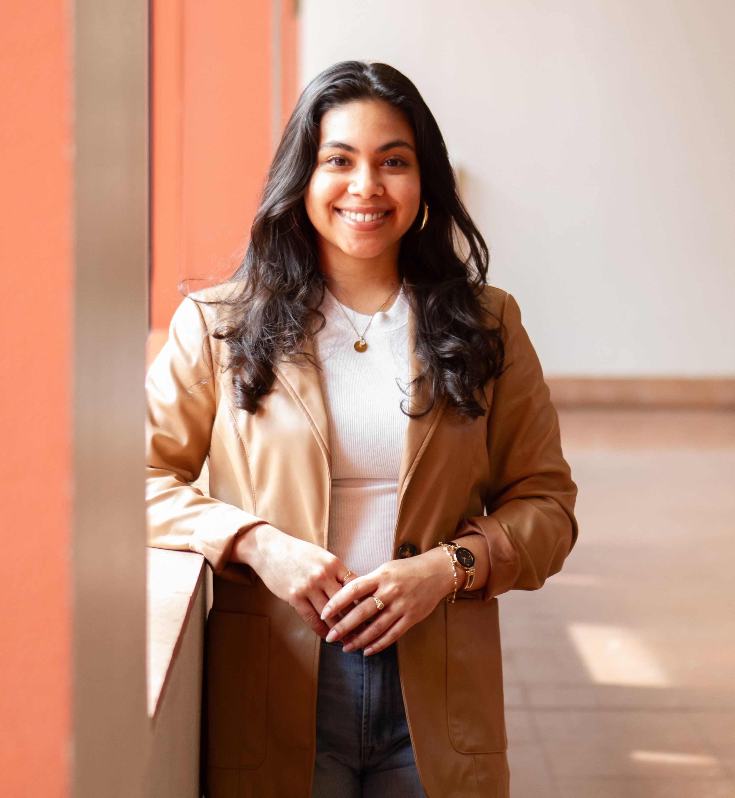 A smiling woman with long, wavy black hair, wearing a tan blazer over a white top, accessorized with gold jewelry, standing indoors near a wall with a reddish-brown color and a beige wall in the background.