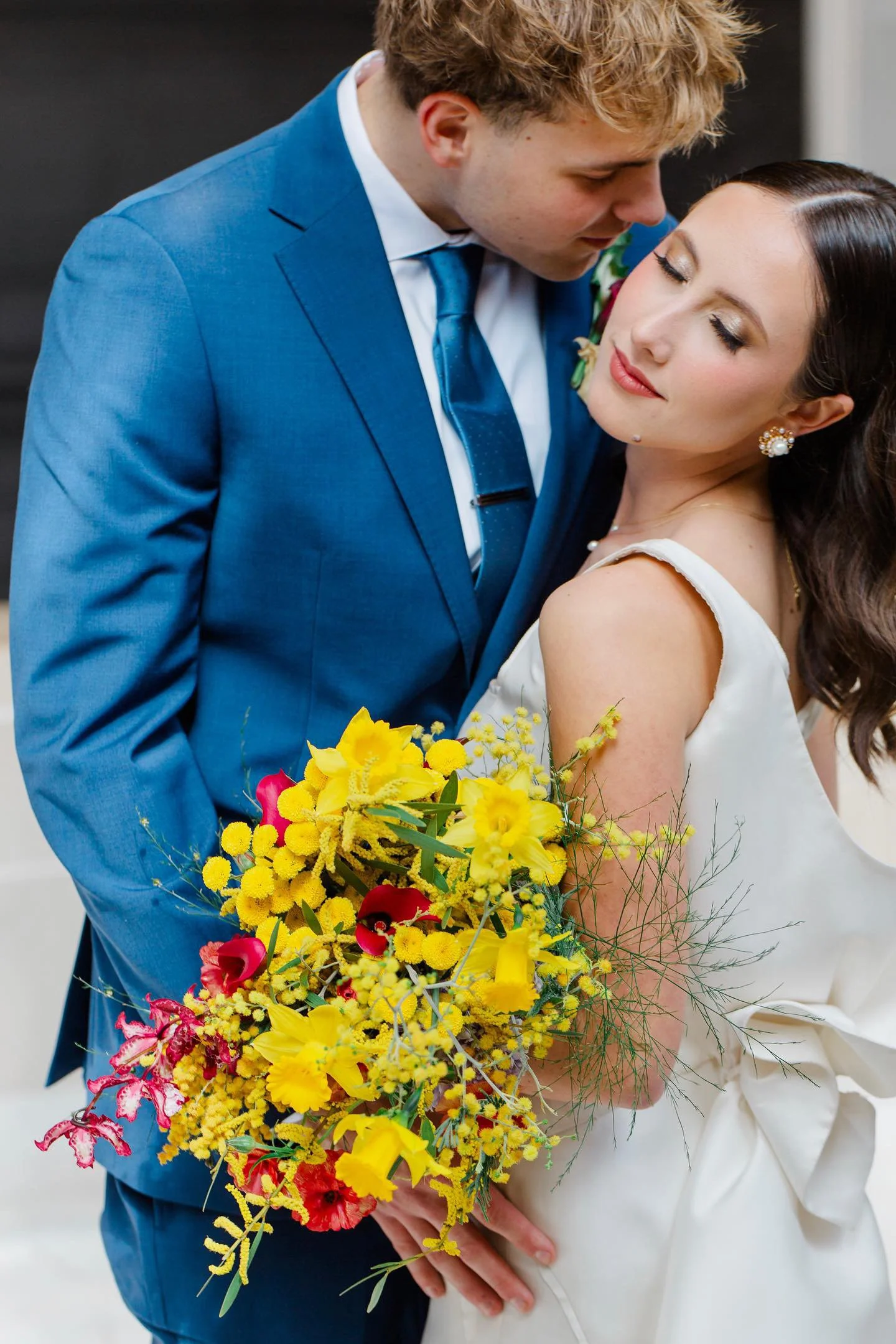 Back view of a woman with styled, curled hair updo and loose curls on sides, wearing a dark plaid shirt, standing against a white brick wall.