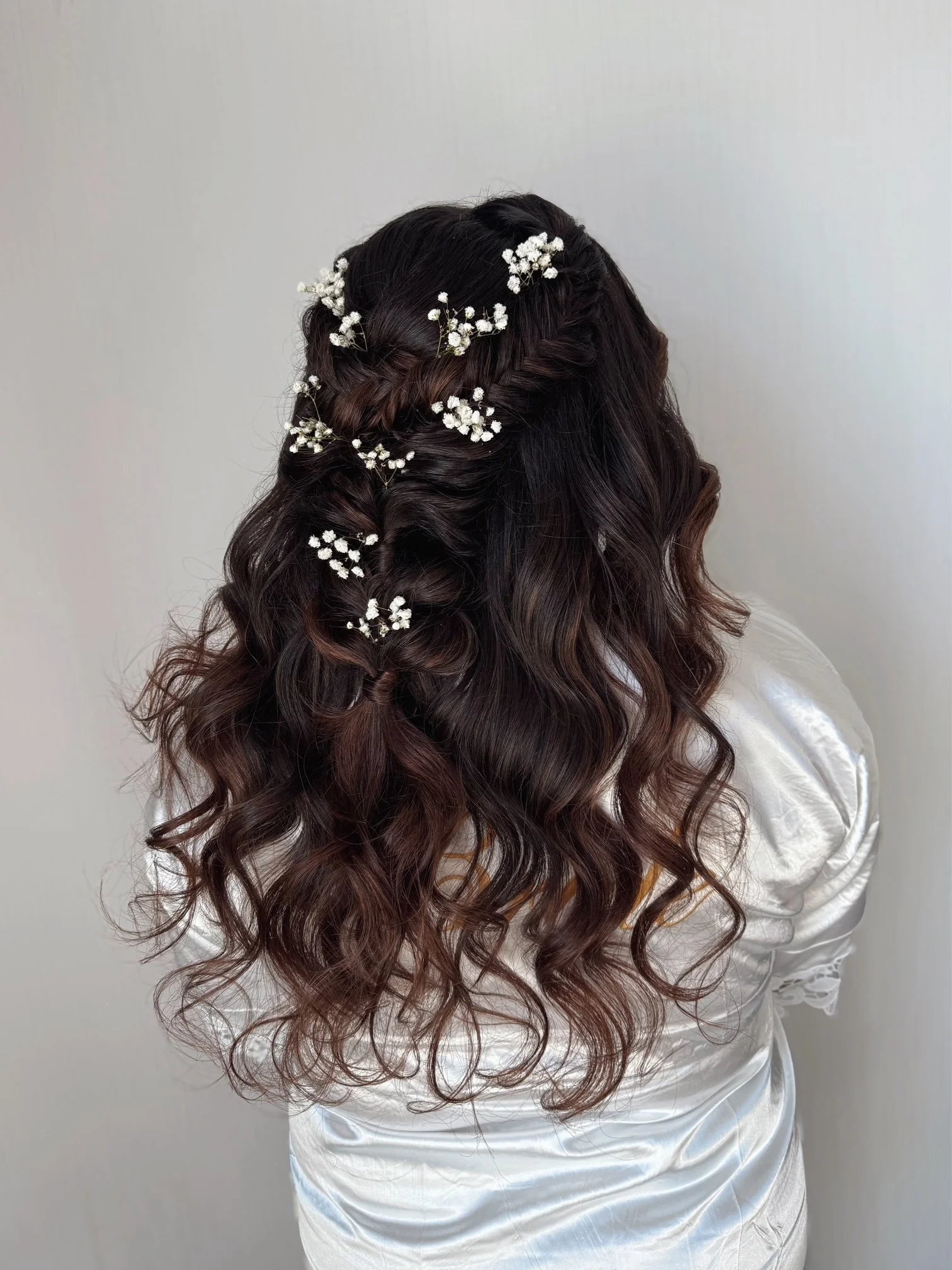 Back view of a woman with long, curly brown hair decorated with small white flower hairpins, wearing a white satin blouse.