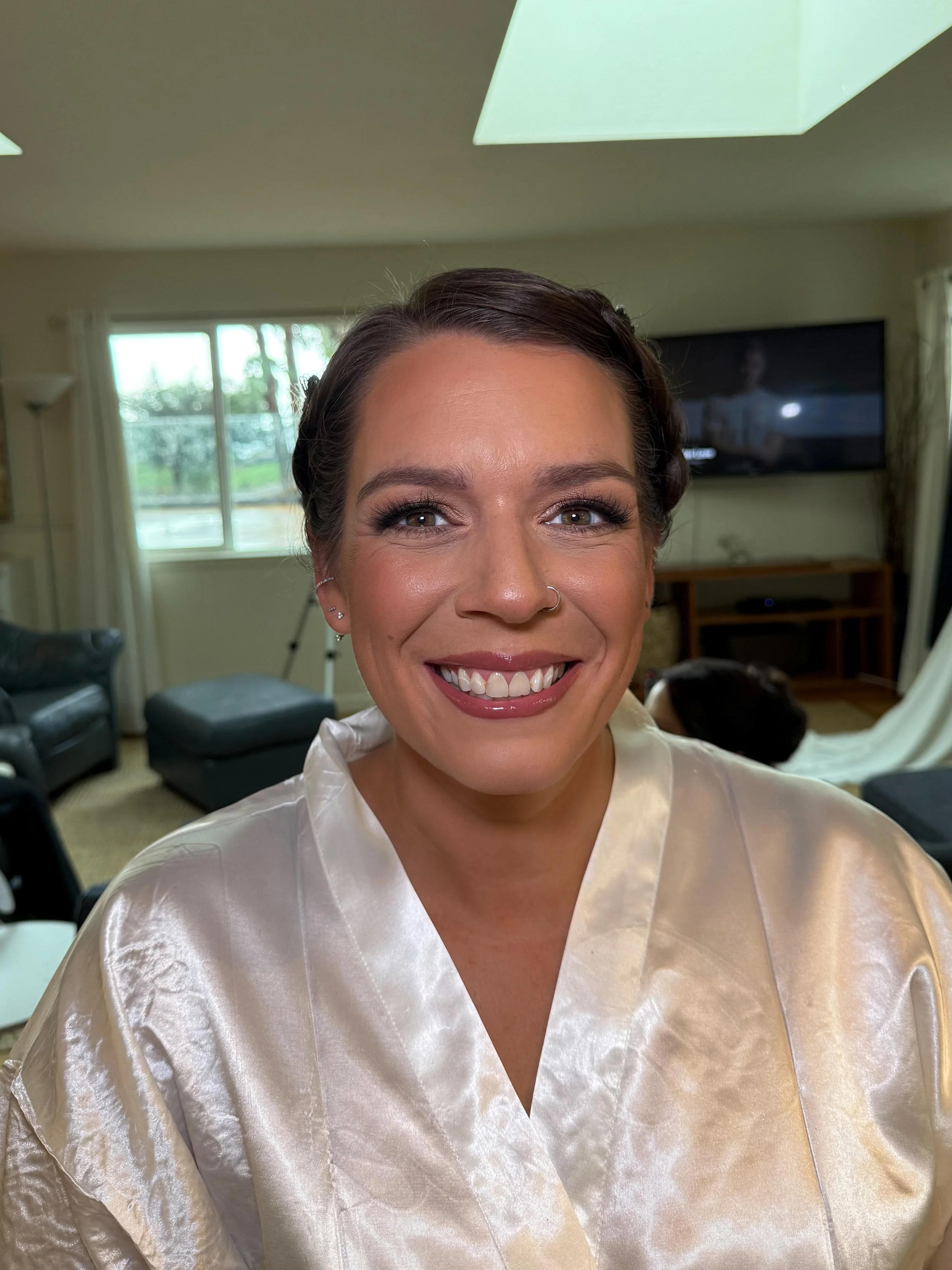 Close-up of a smiling woman with makeup and a nose ring, wearing a satin robe, in a living room with a TV, curtains, and a dog in the background.