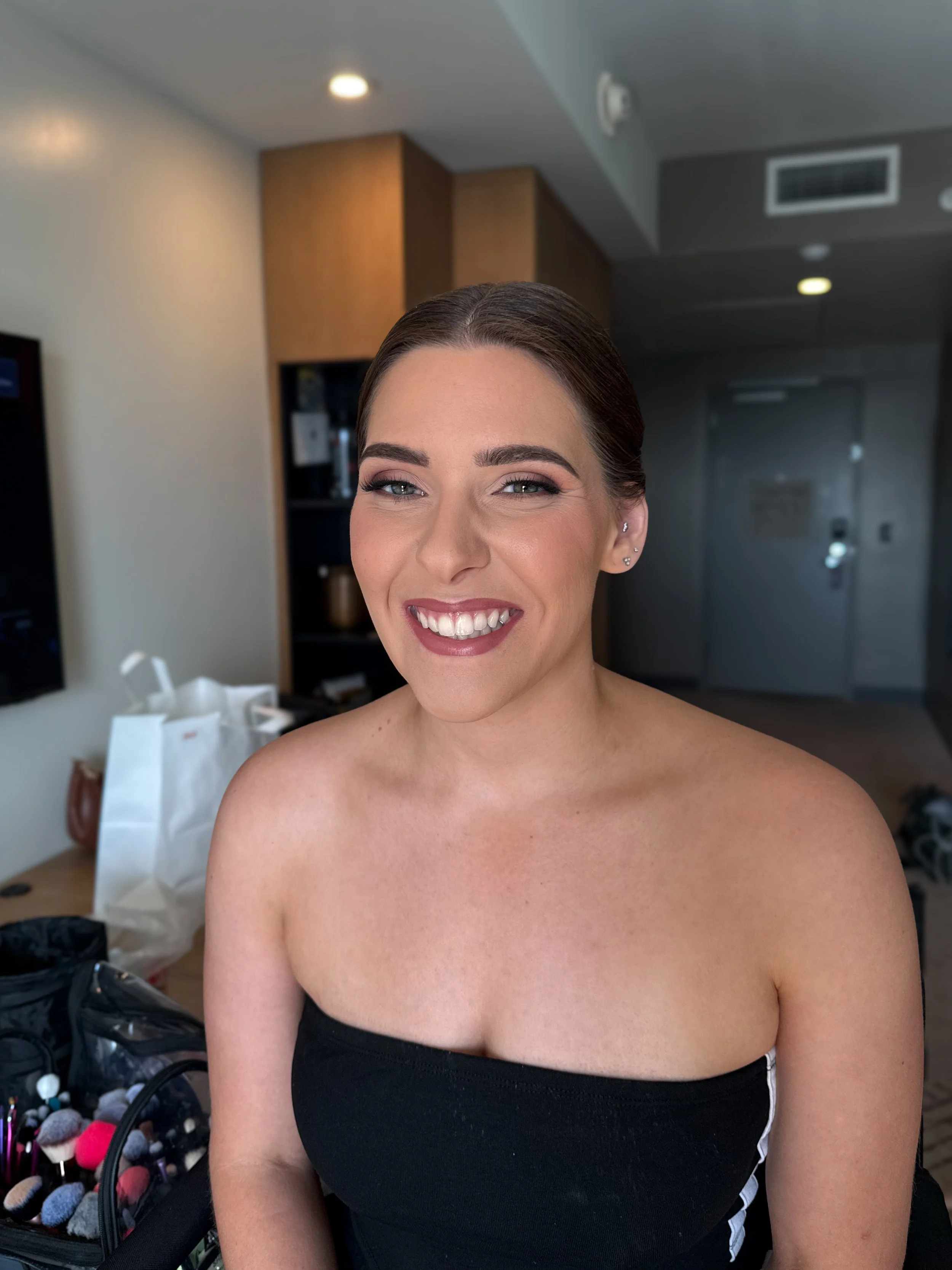 A smiling woman with makeup in a hotel room, wearing a strapless black top.
