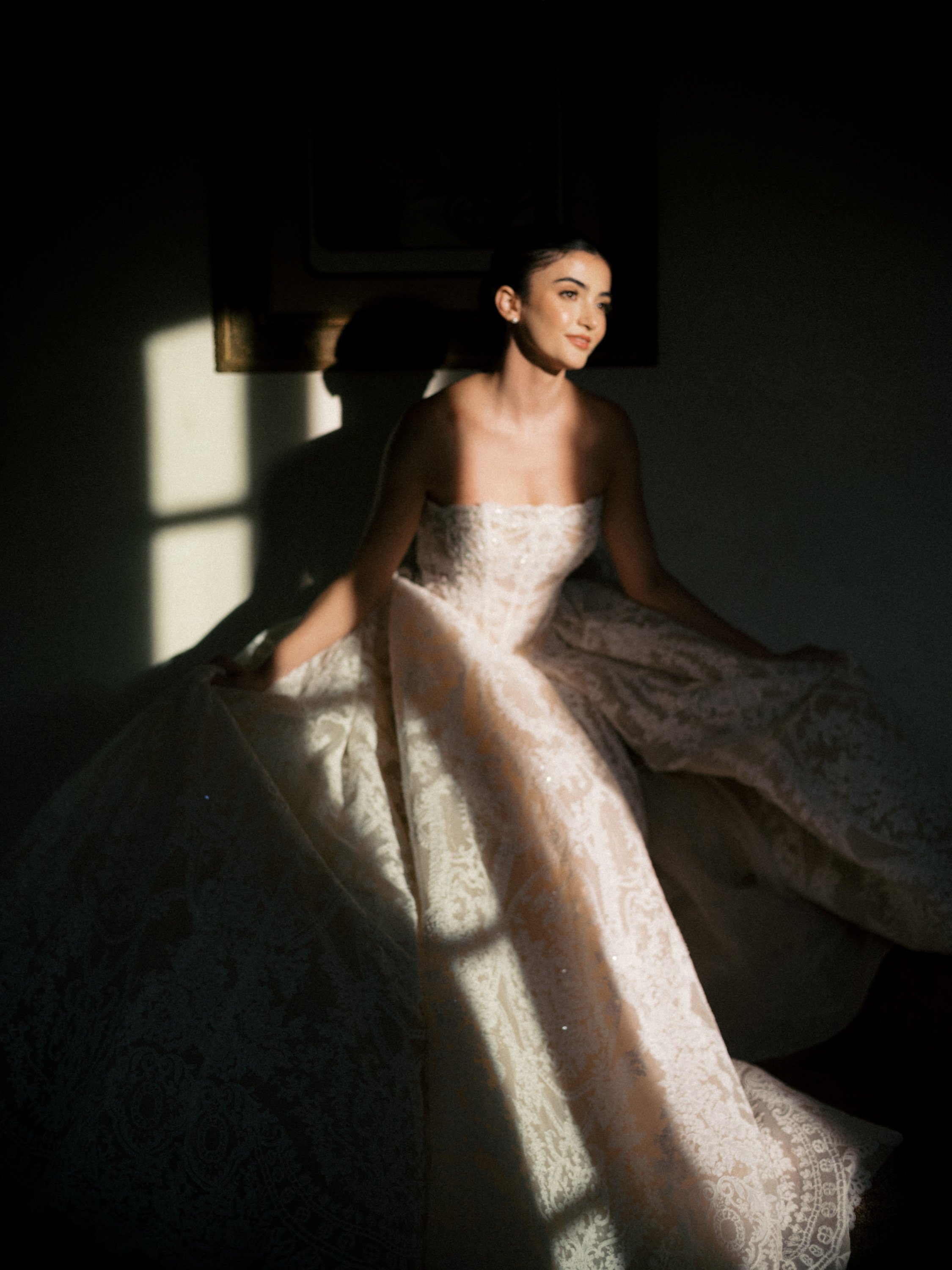 Back view of a woman with styled, curled hair updo and loose curls on sides, wearing a dark plaid shirt, standing against a white brick wall.