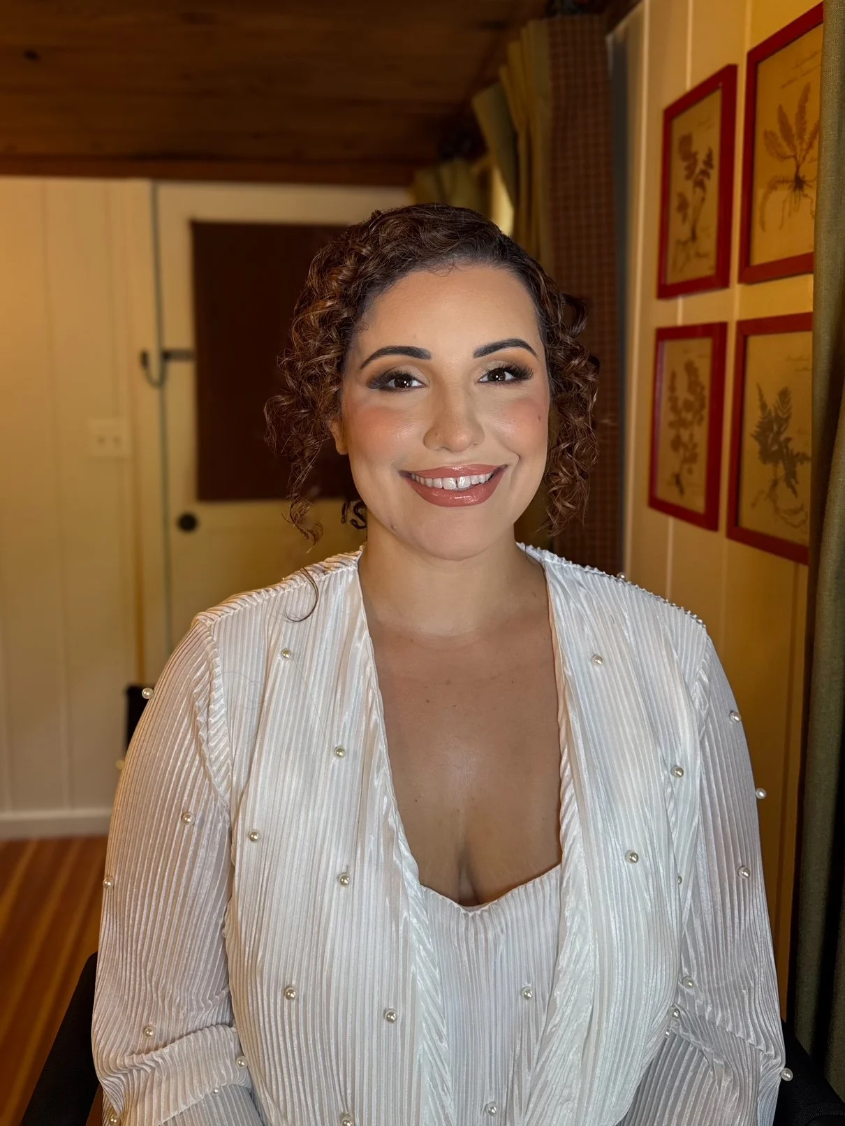 A woman with curly brown hair and makeup, smiling, wearing a white pleated chiffon dress with pearl embellishments, sitting in a room with yellow walls and framed botanical artwork.