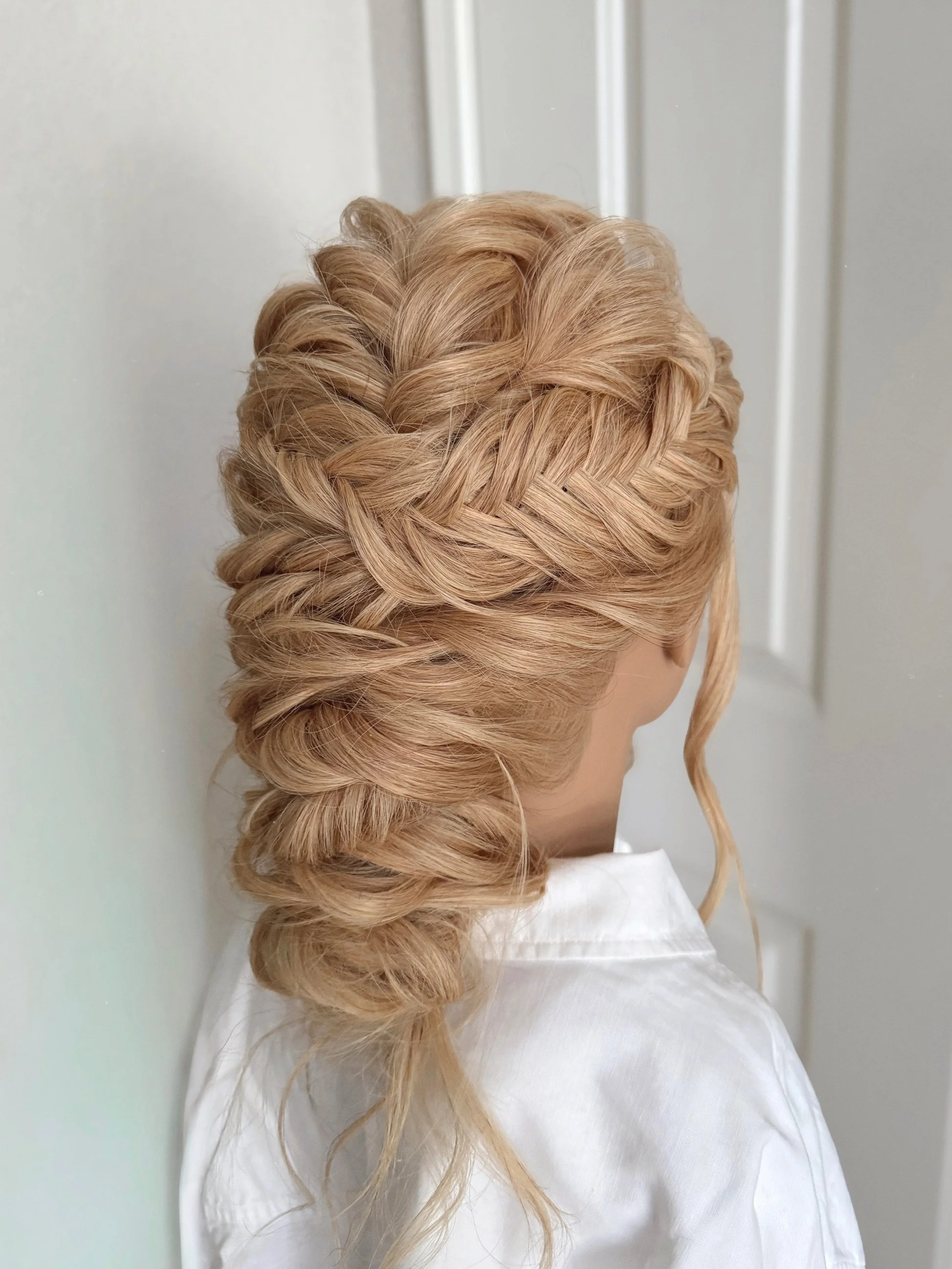 A woman with elaborate blonde braided hairstyle, with her hair styled in a crown and cascading into an elegant loose braid, wearing a white blouse.