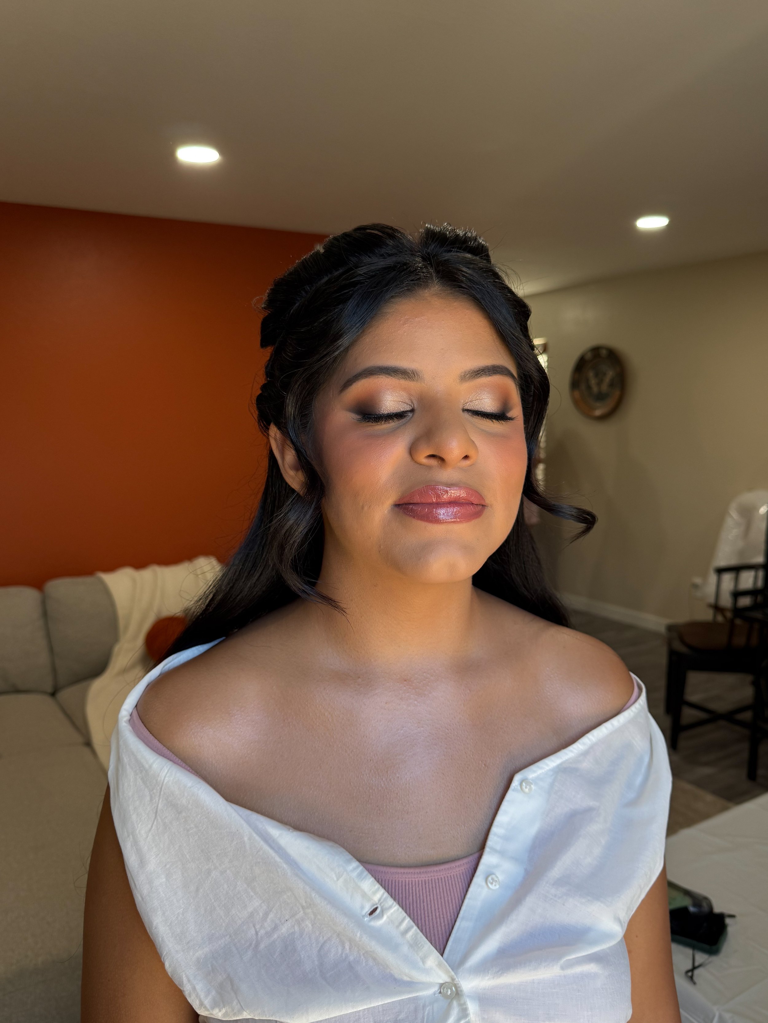 A woman with closed eyes, wearing makeup, with long dark hair, and a white shirt off the shoulders, standing in a room with a beige and orange wall.