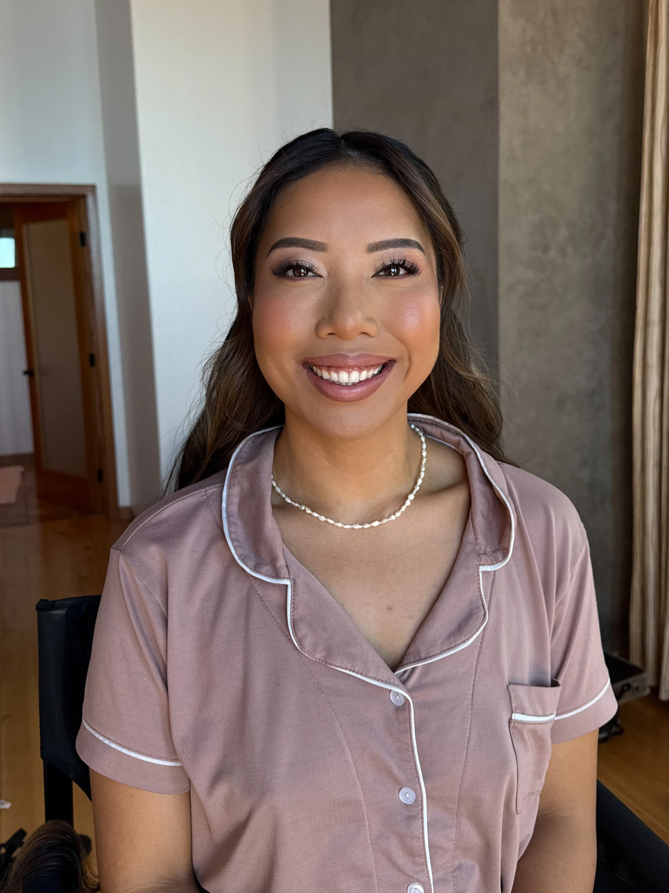 A young woman smiling, wearing a light pink satin pajama top and a pearl necklace in an indoor setting.