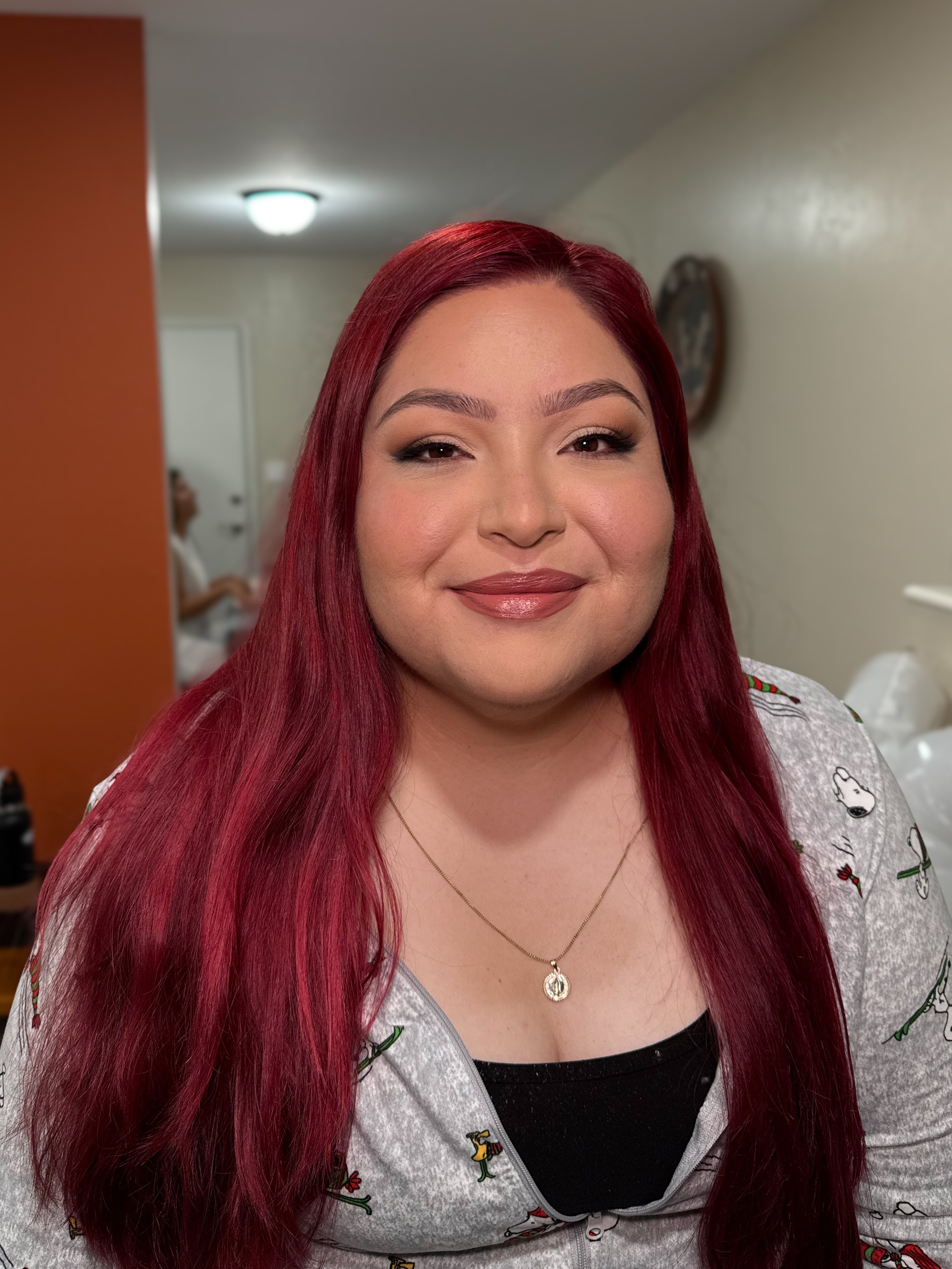 A woman with long red hair and makeup, wearing a gray top with cartoon characters, and a gold necklace, smiling at the camera indoors.