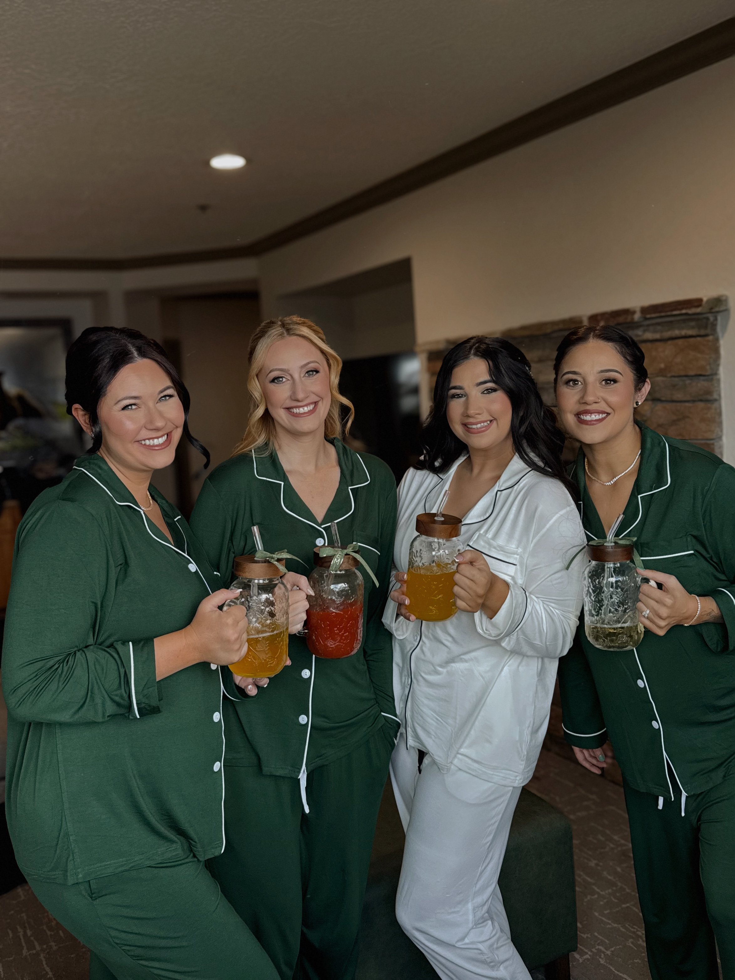 Four women dressed in pajamas, smiling, holding mason jar mugs of drinks, standing indoors with a brick fireplace in the background.