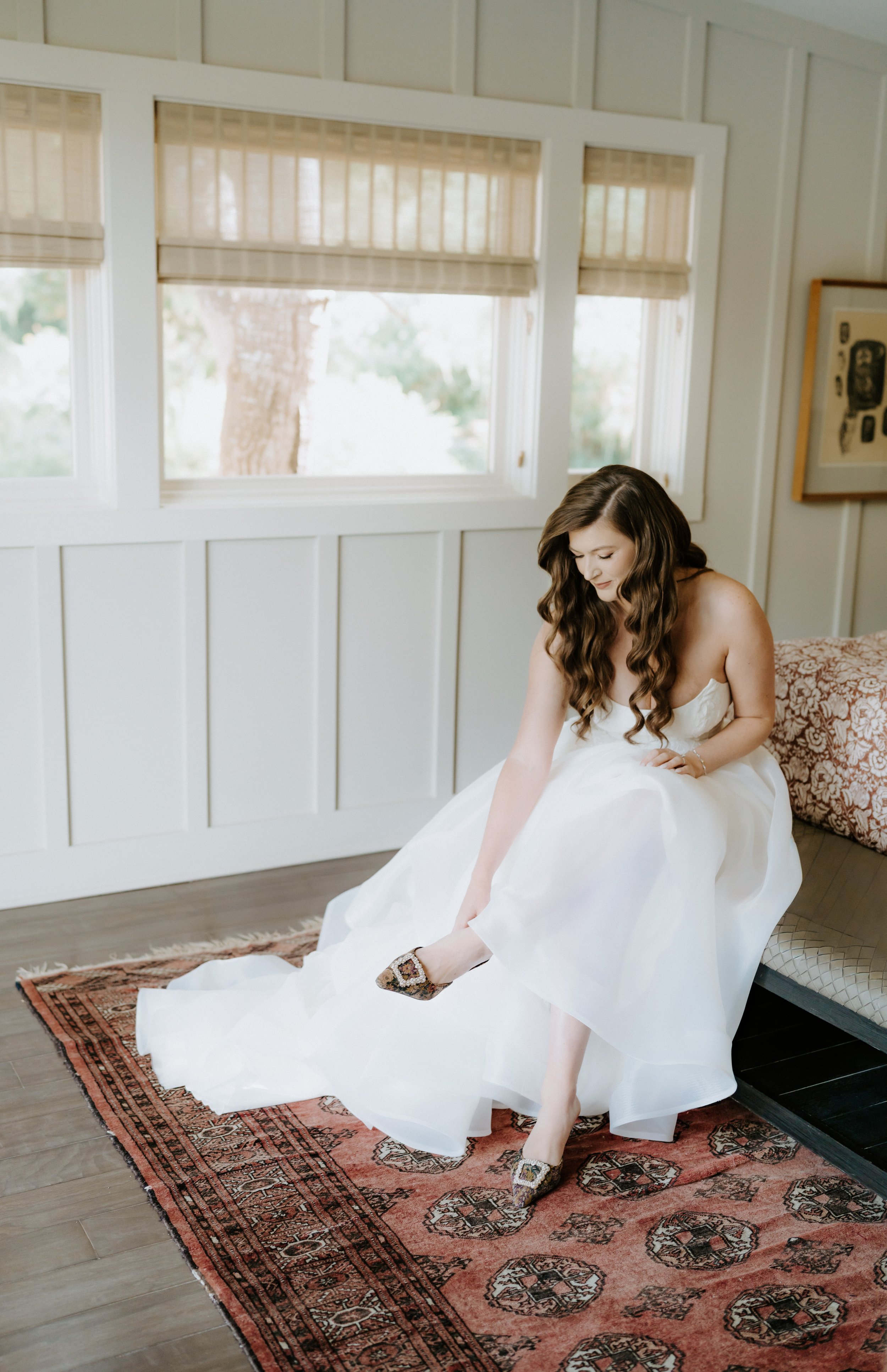 A woman in a white wedding dress sitting on a bench, adjusting her shoe while indoors with large windows and a patterned rug.
