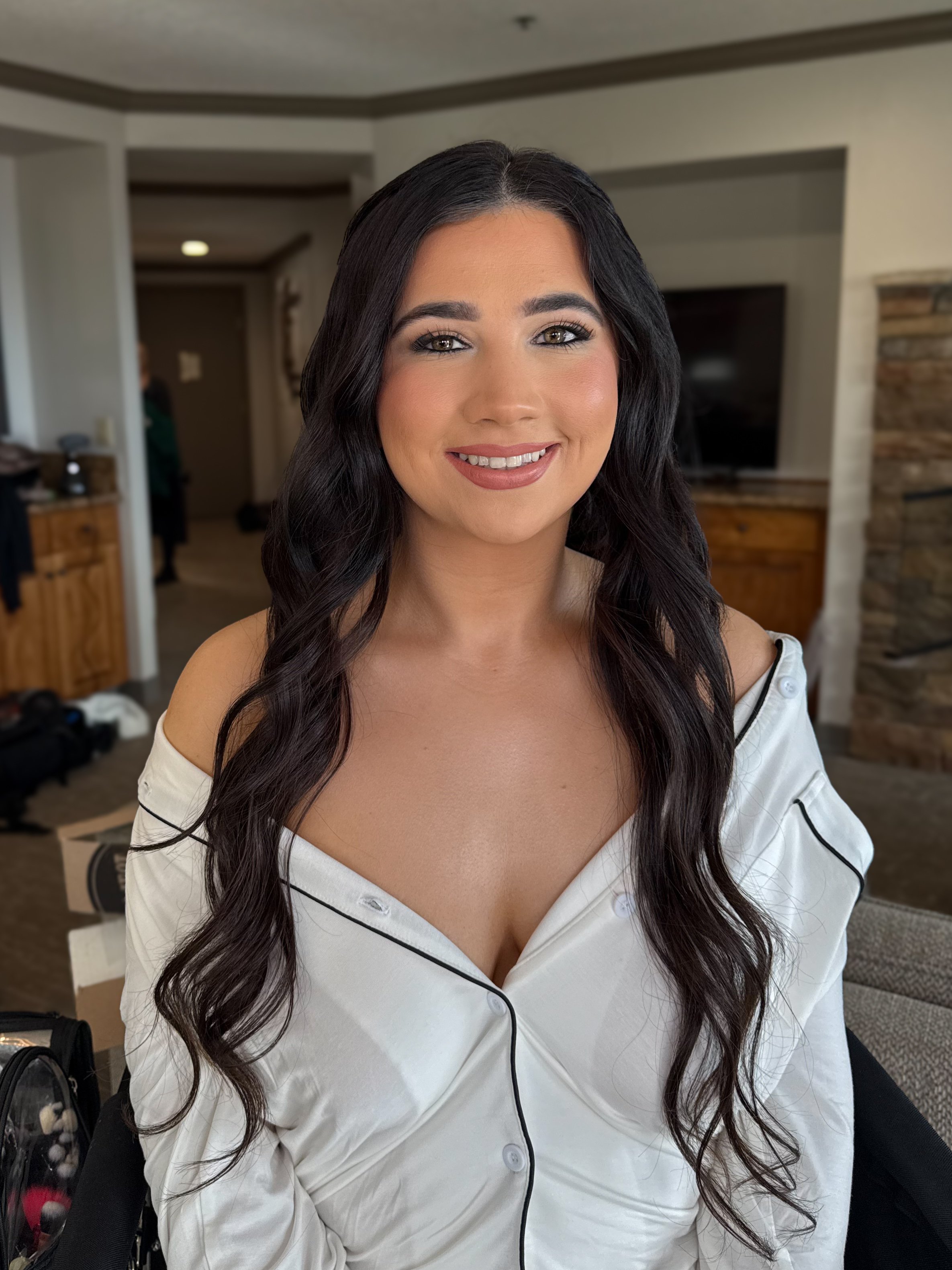 A young woman with long, dark wavy hair, smiling, wearing an off-the-shoulder white satin shirt, indoors with a blurred background of a living room.