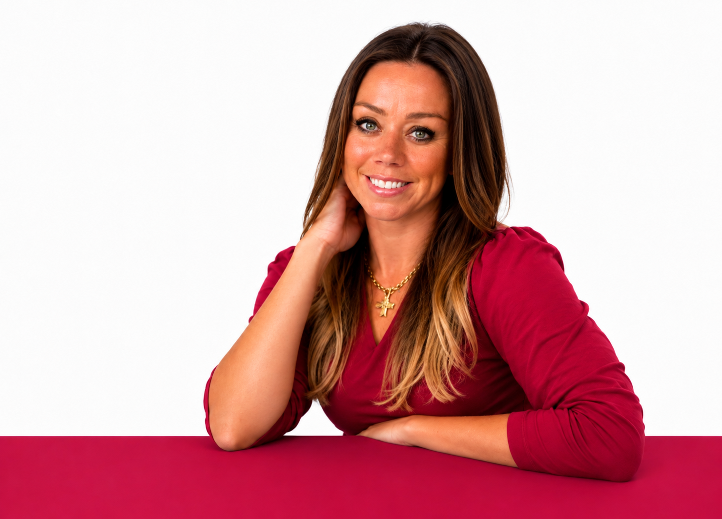 A woman with long brown hair, wearing a red top and a gold necklace, sitting at a red table, smiling at the camera against a white background.