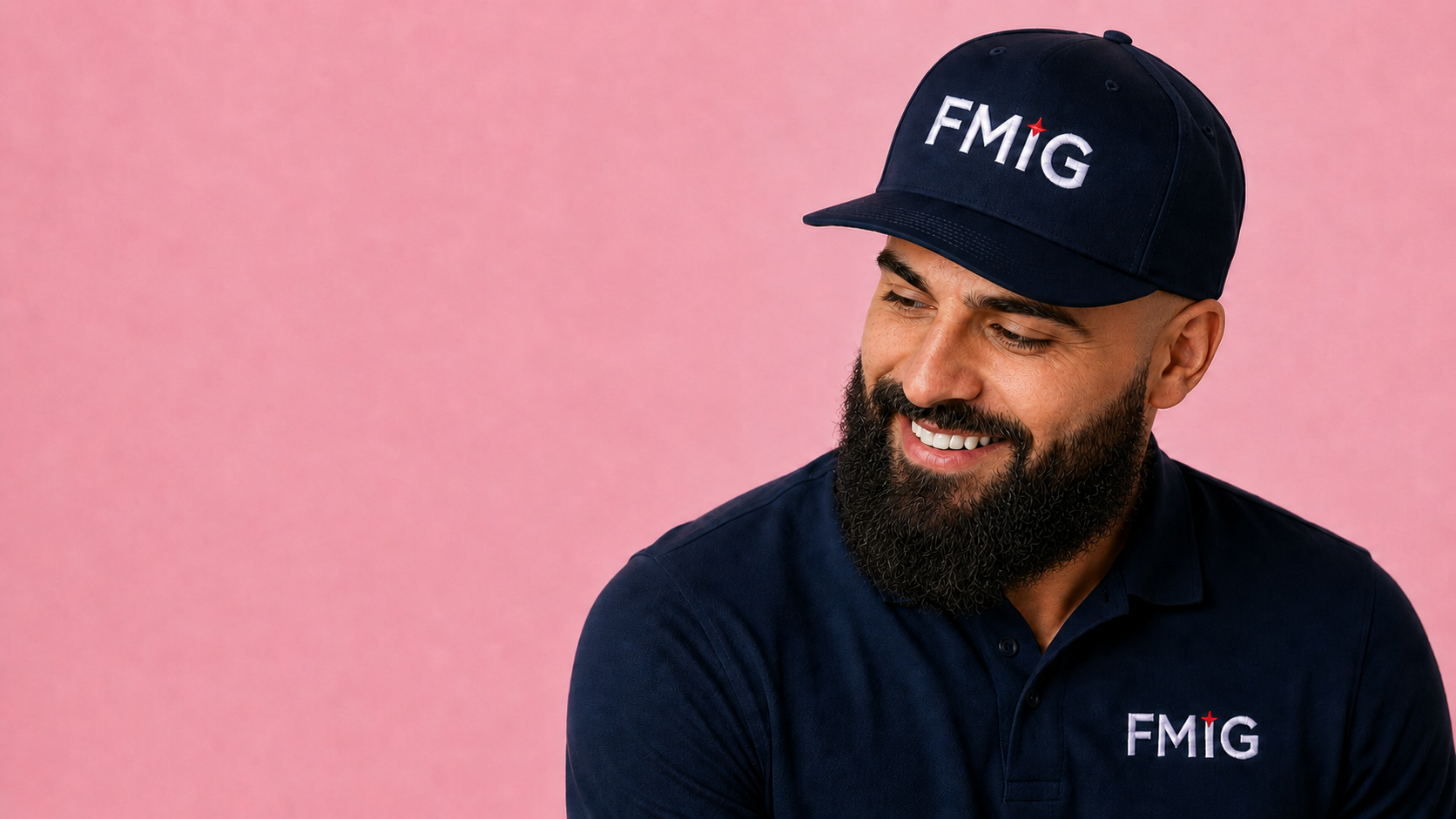A man with a beard wearing a navy blue cap and shirt, both with the FMIG logo, smiling against a pink background.