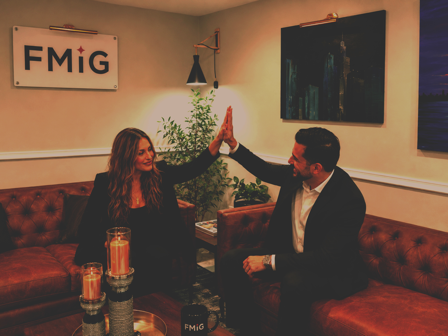 A man and woman sitting on a leather couch giving each other a high five indoors. The woman has long wavy hair and is wearing a black blazer, while the man is wearing a dark suit and white shirt. There are candles on a table, plants, and paintings on the wall, with a sign that reads 'FMIG' in the background.