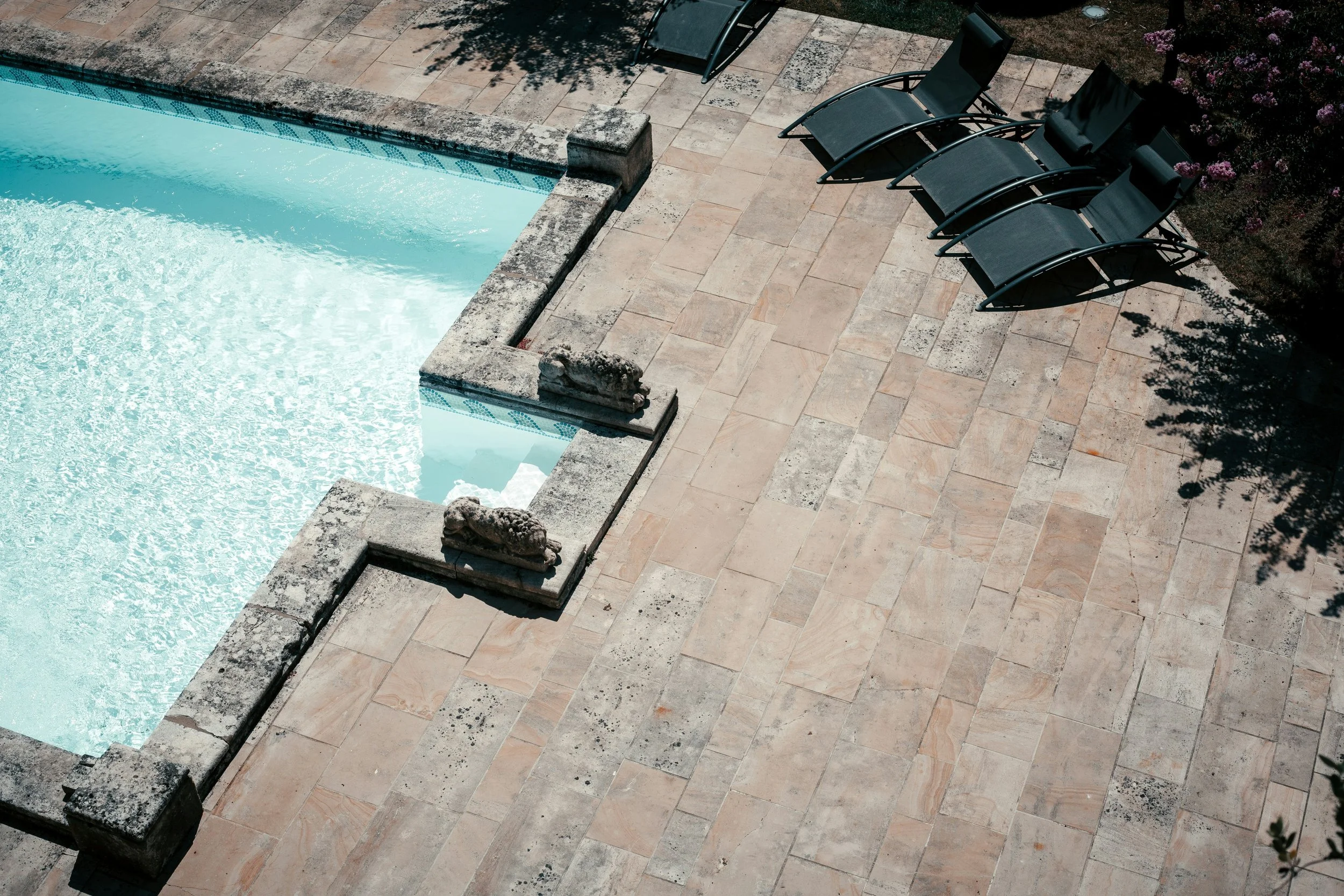An outdoor swimming pool with light blue water and a weathered stone border, surrounded by a stone patio with lounge chairs and garden foliage.