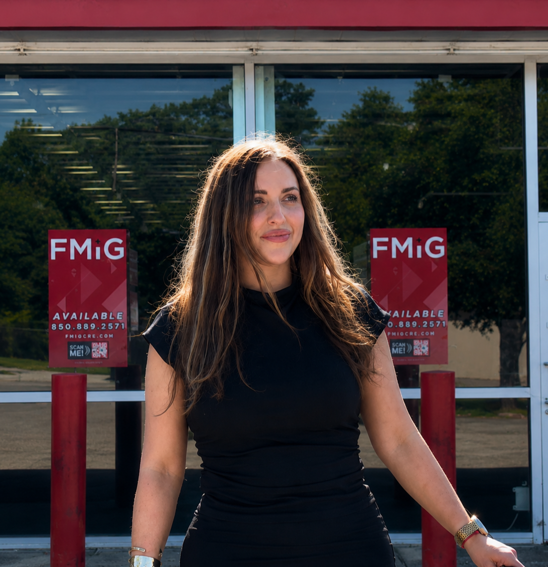 A woman with brown hair in a black top standing outside a building with FMIG signs in the background.