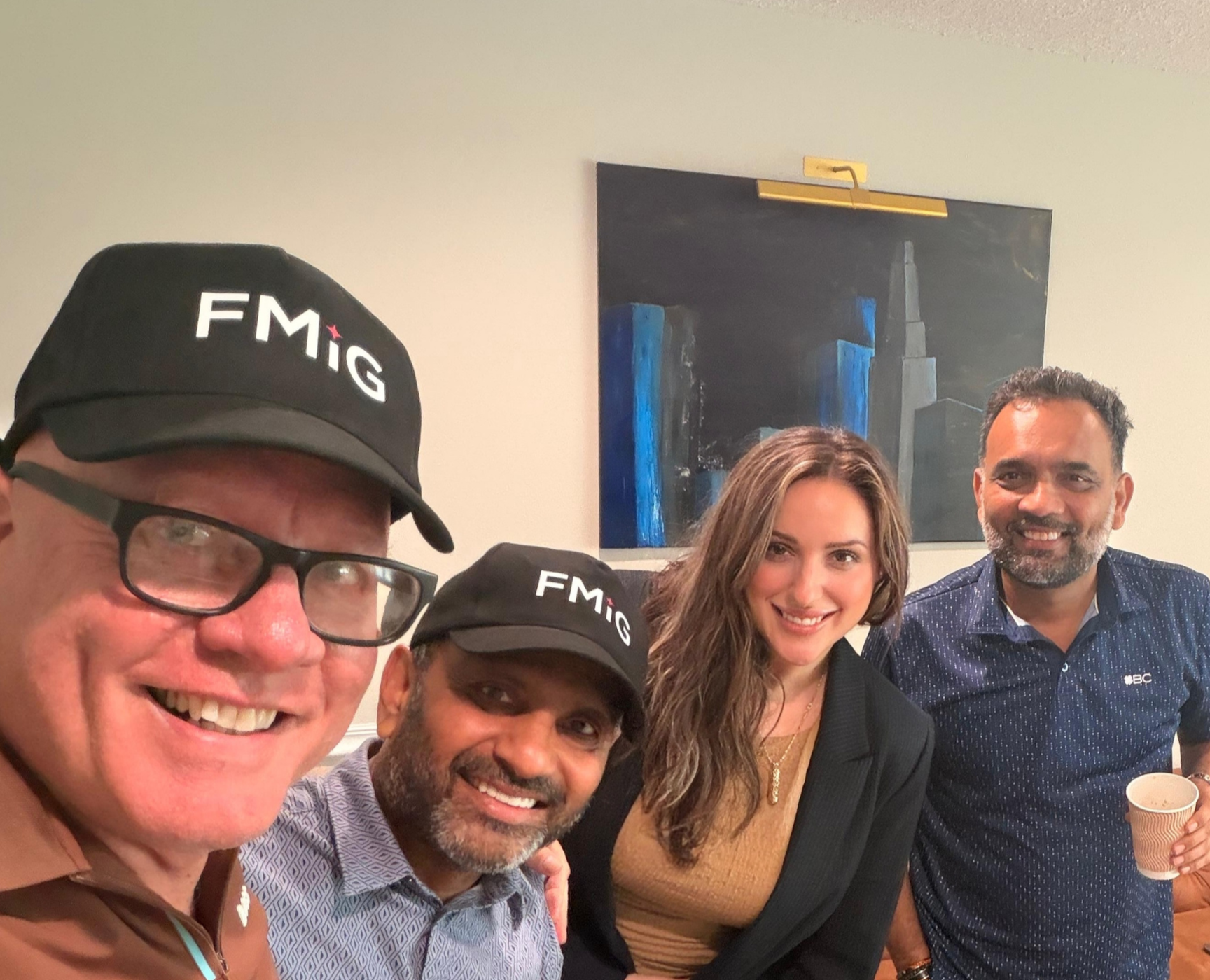 Group of five people smiling at a social gathering, two wearing black caps with 'FMIG' printed on them. A man wearing glasses and a brown shirt, a woman with long wavy hair and a black blazer, and another man in a navy blue patterned shirt holding a paper cup are visible, with a woman in a gold top at the center and a man with a beard to the right. There is a painting of a city skyline on the wall behind them.